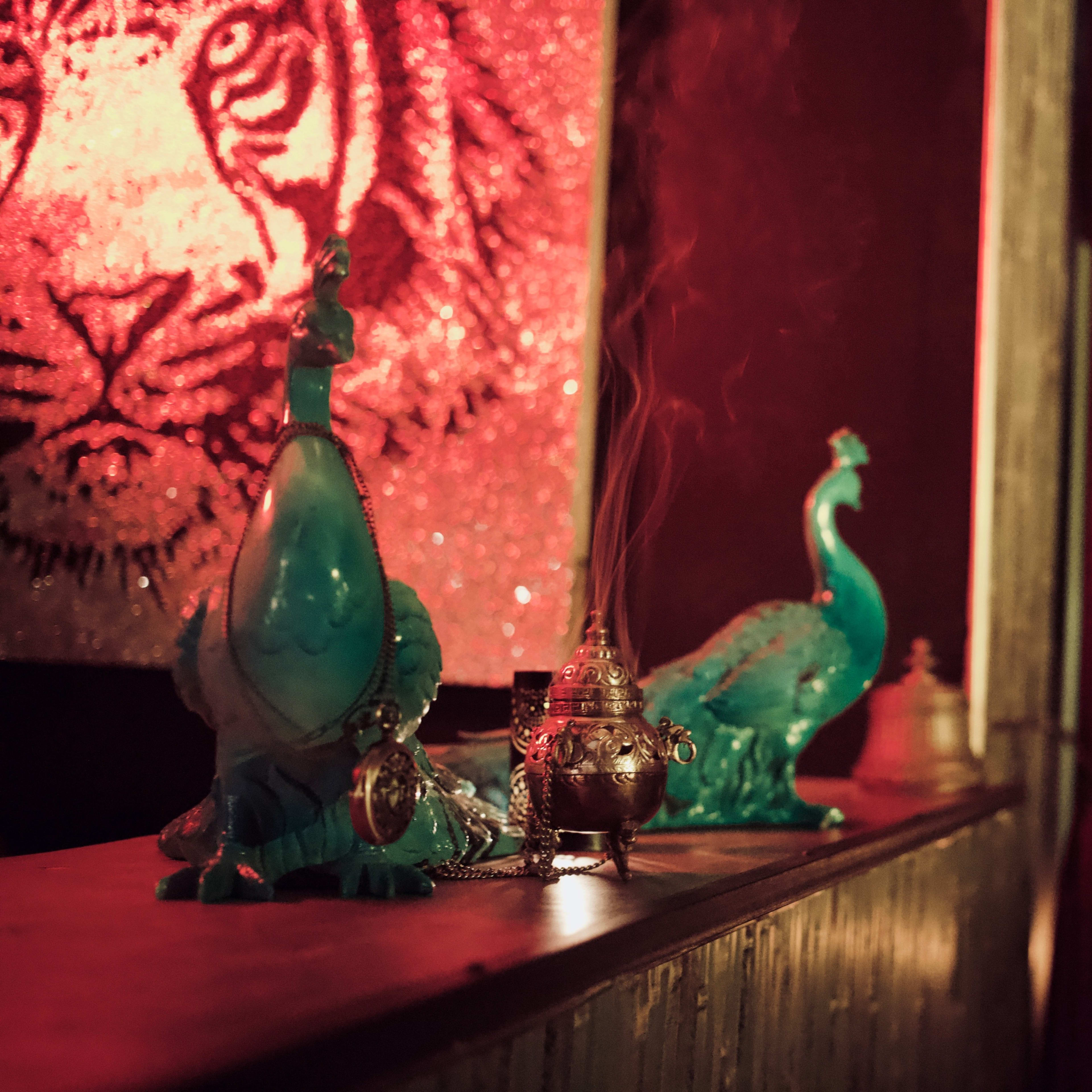 The scene features two turquoise peacock figurines and a small ornate incense burner on a wooden ledge, with a vibrant red backdrop illuminated by soft lighting.