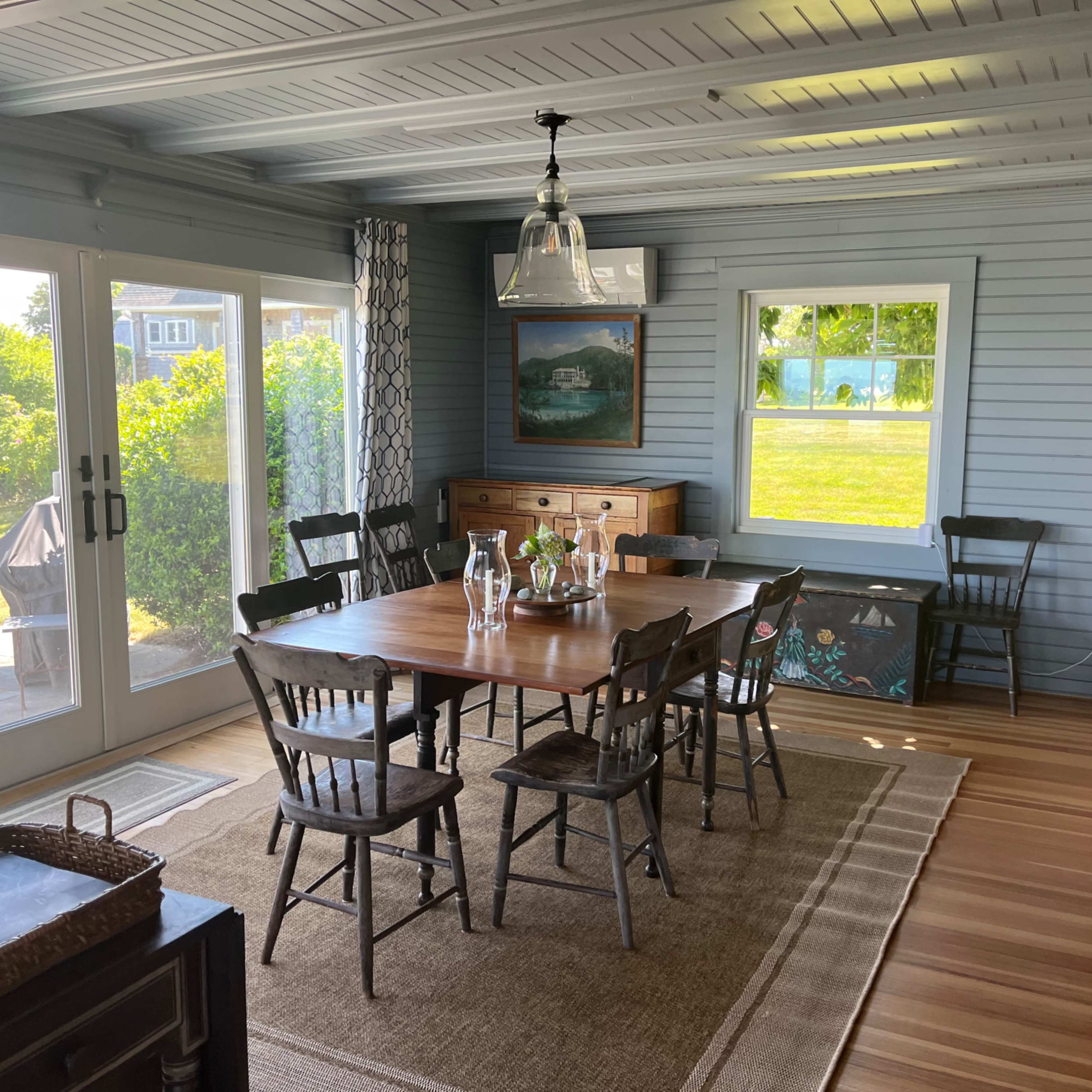 A sunlit dining area with a large wooden table surrounded by six black chairs, a sideboard, and a cupboard against the walls of a blue-paneled room.