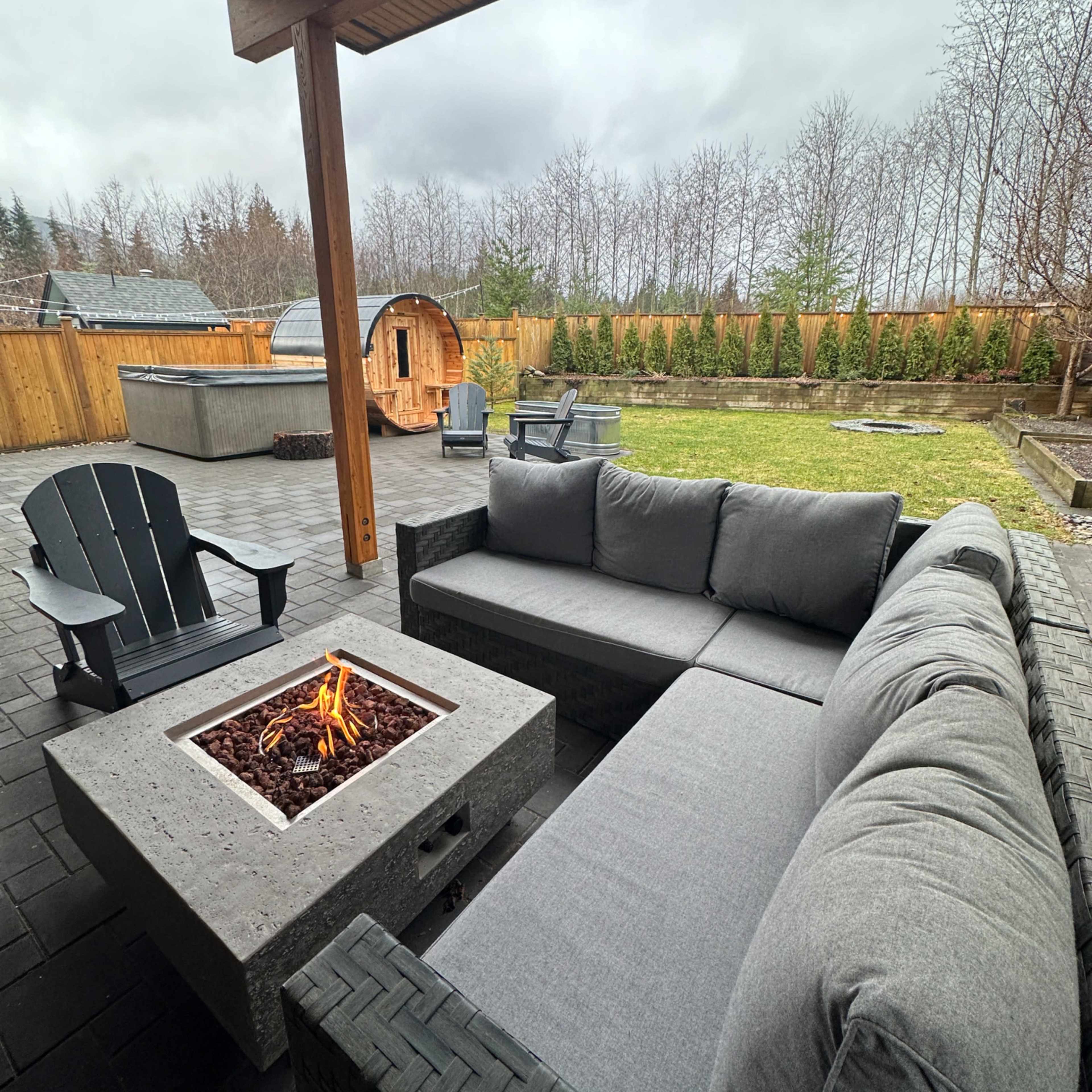 A modern outdoor seating area features a gray sectional sofa and a square fire pit, surrounded by a grassy yard and wooden fencing under a cloudy sky.