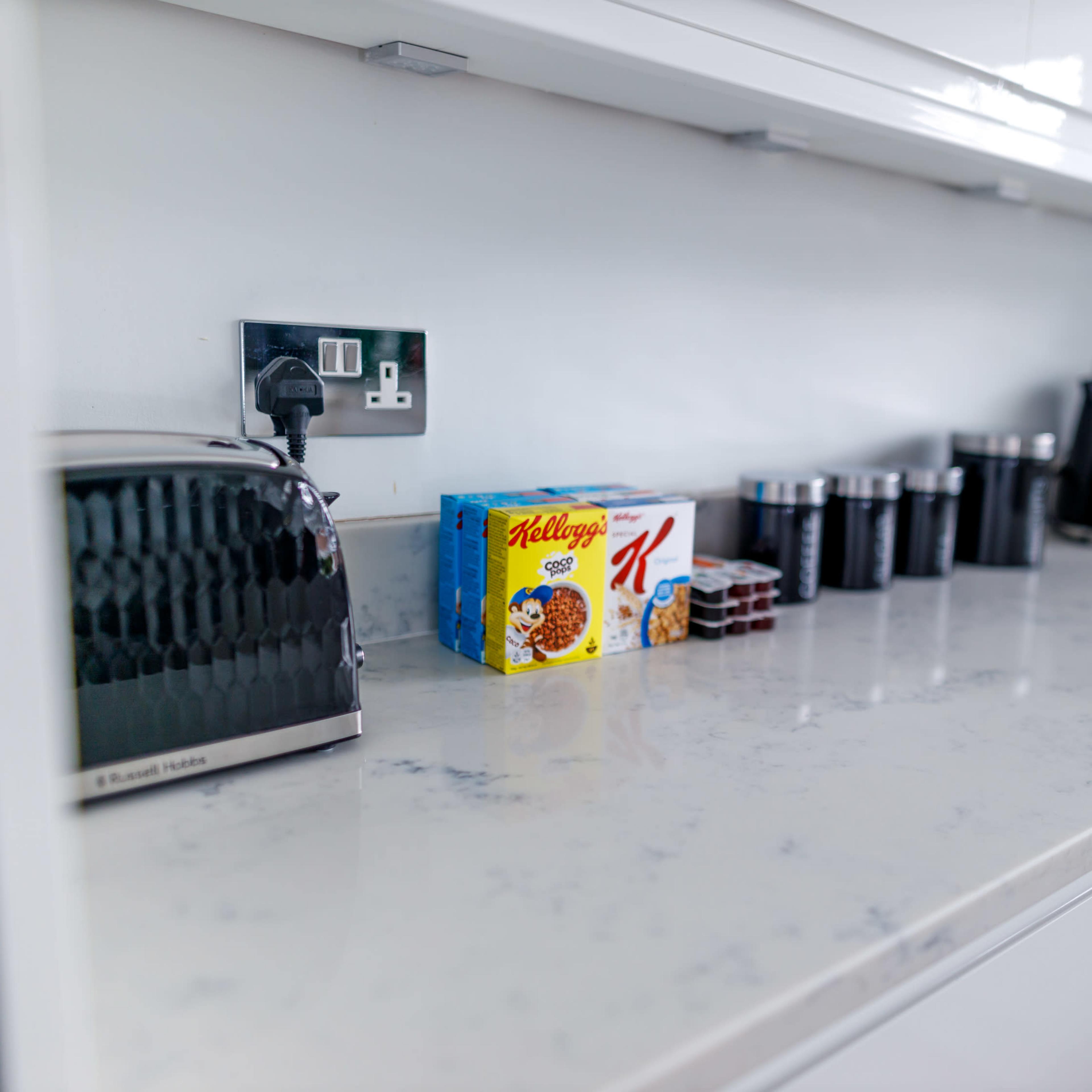 A modern kitchen countertop features a toaster, a box of cereal, jars, and kitchen utensils neatly arranged against a white backdrop.