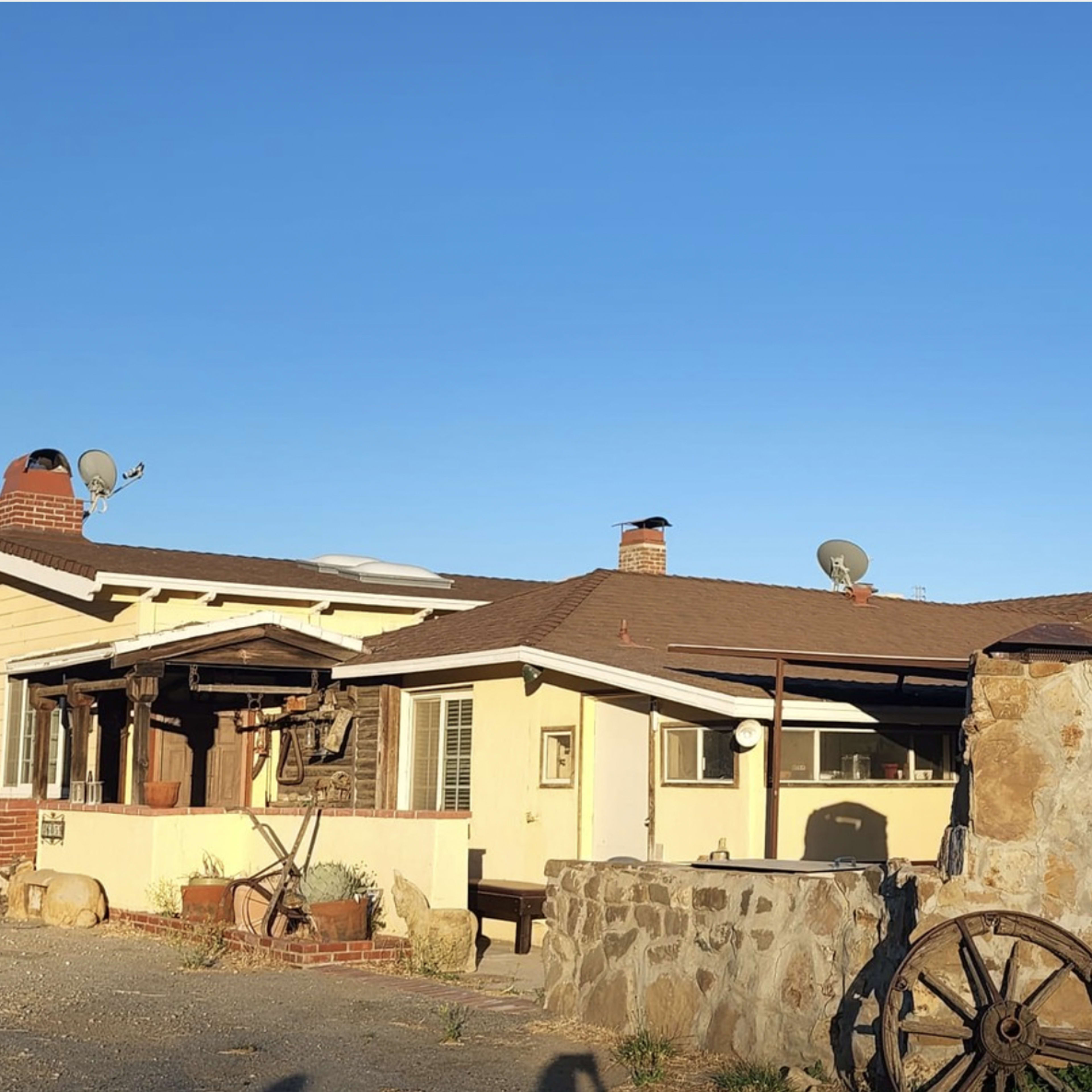 A yellow rural house with a stone wall and an old wooden cart wheel is set against a clear blue sky and hills in the background.