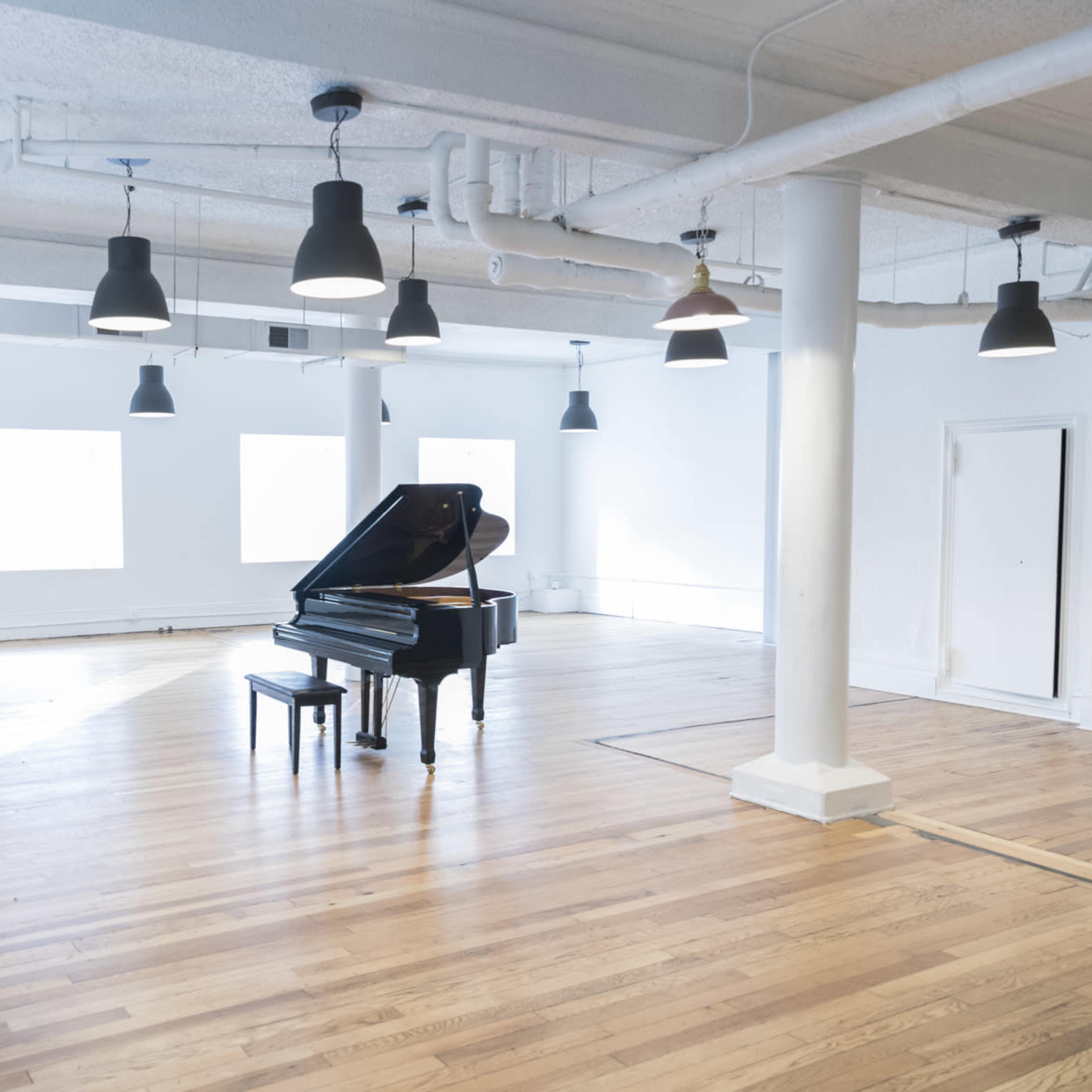 A grand piano sits alone in a spacious, brightly lit room with wooden floors and large windows.
