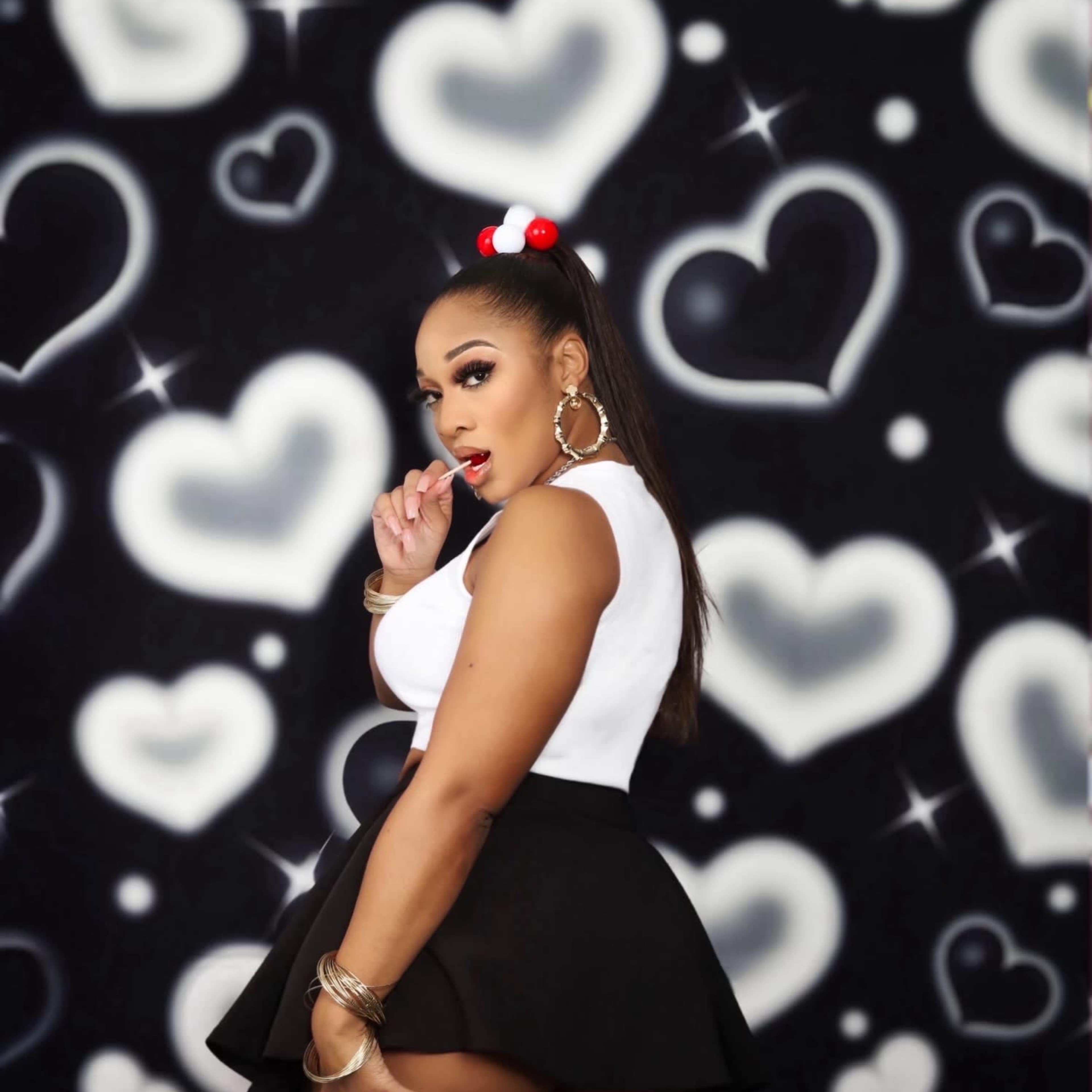 A woman in a white top and black skirt poses playfully against a backdrop of heart shapes.