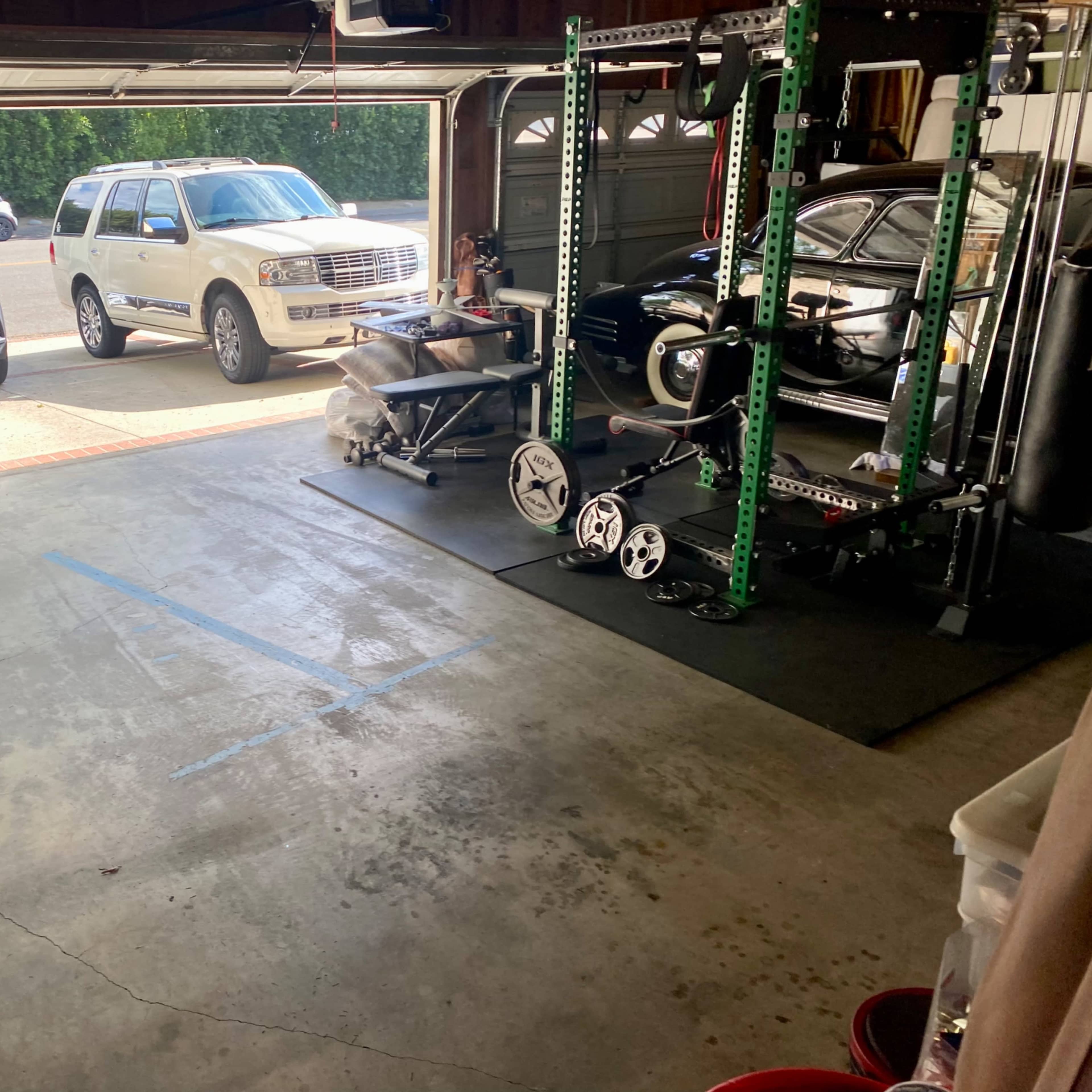 A garage with workout equipment on one side and two parked vehicles outside, including an SUV and a sedan.