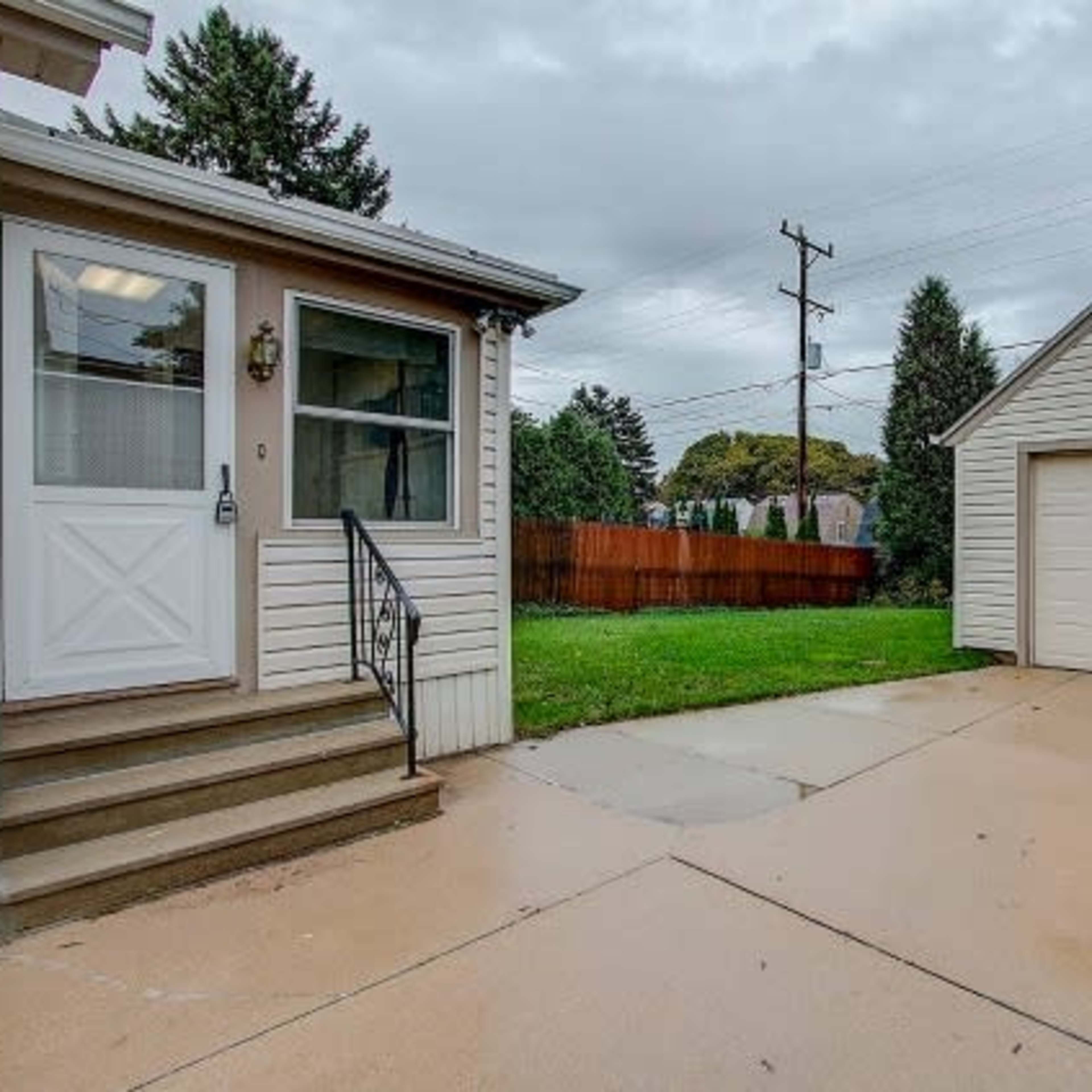 A house entrance with a concrete pathway leading to a beige garage in a grassy area, surrounded by trees and a wooden fence.