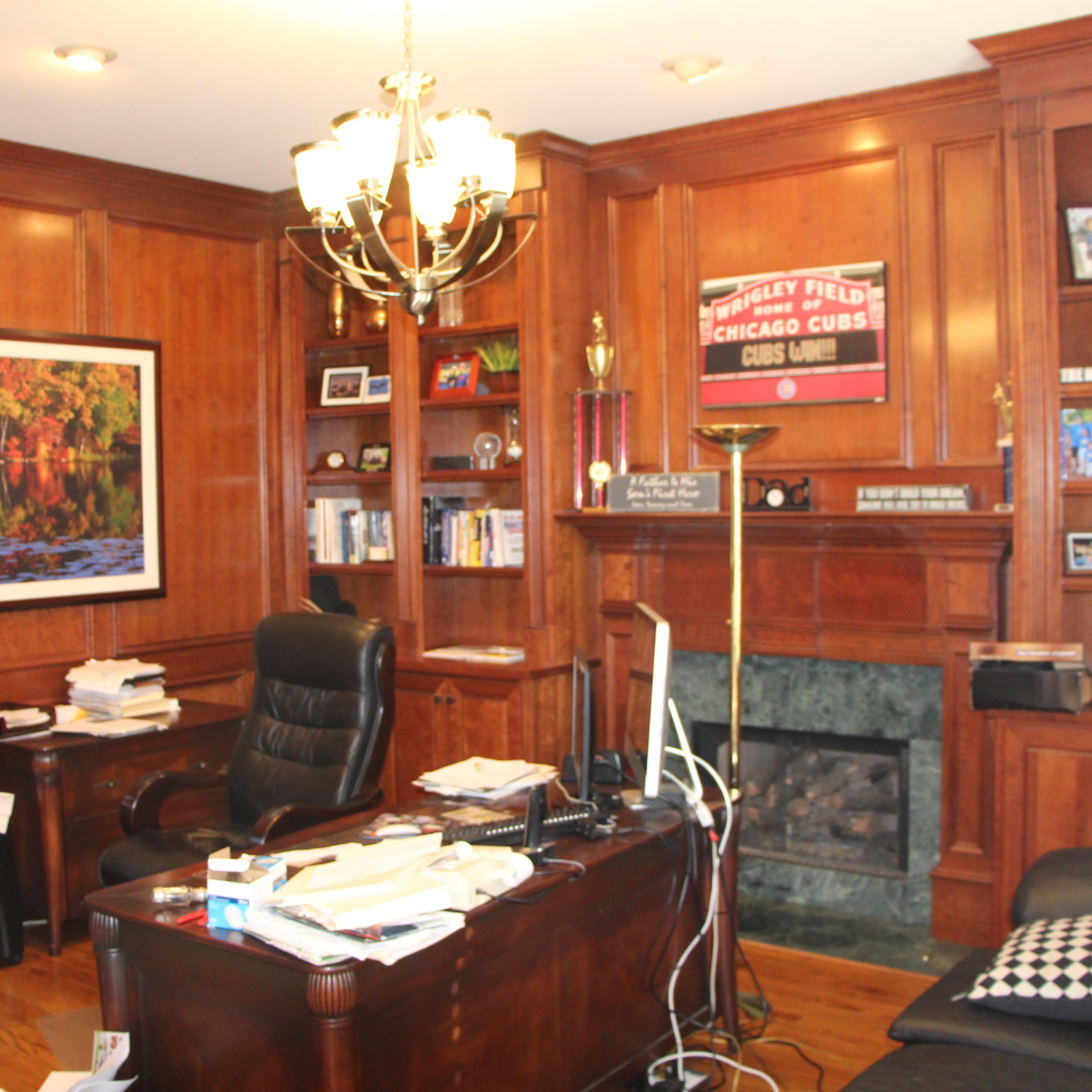 A wooden-paneled office with a desk, black leather chair, bookshelves, and a large framed photo of a scenic landscape on the wall.