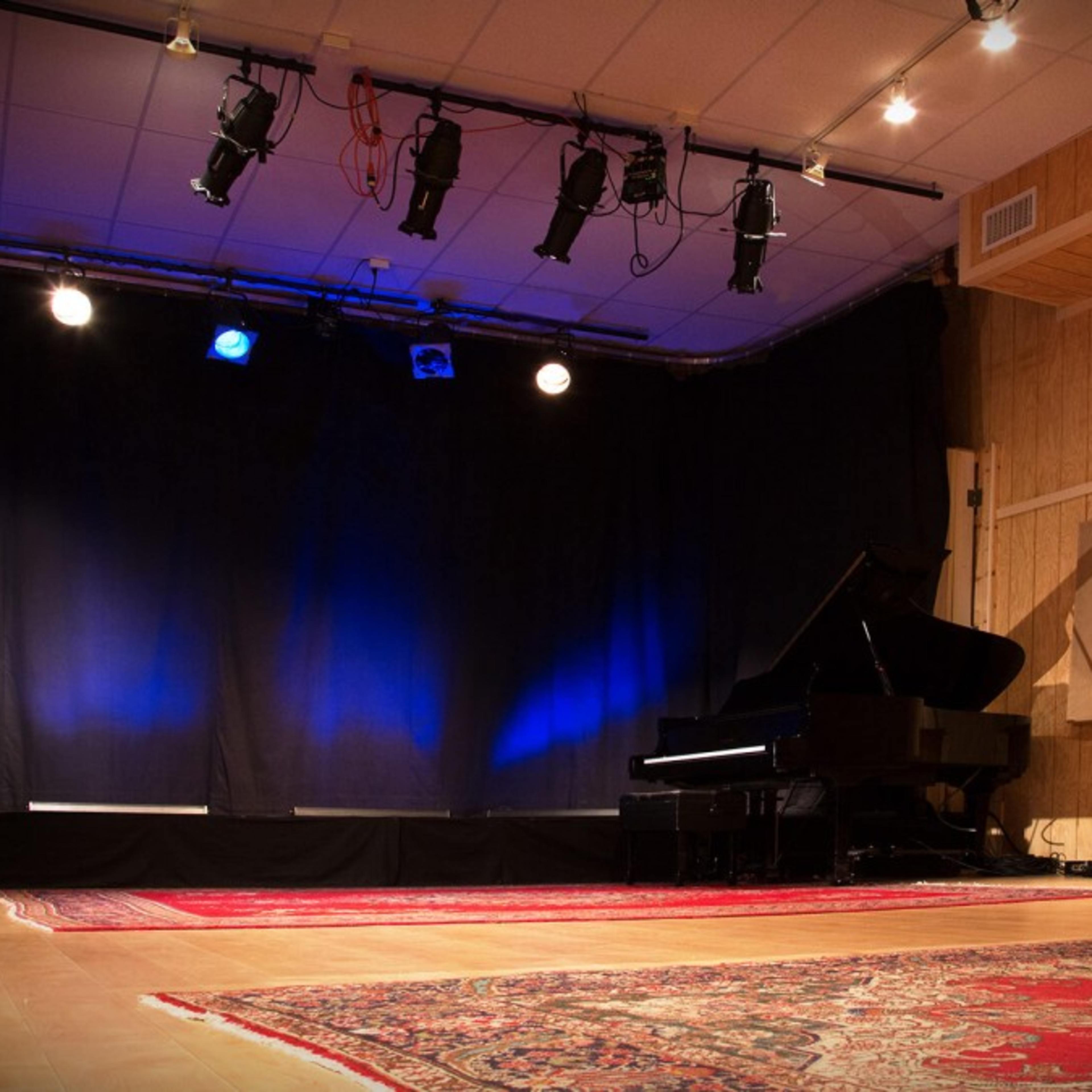 A sparsely decorated stage with a grand piano and blue lighting, set in a room with wooden paneling and soundproofing panels.