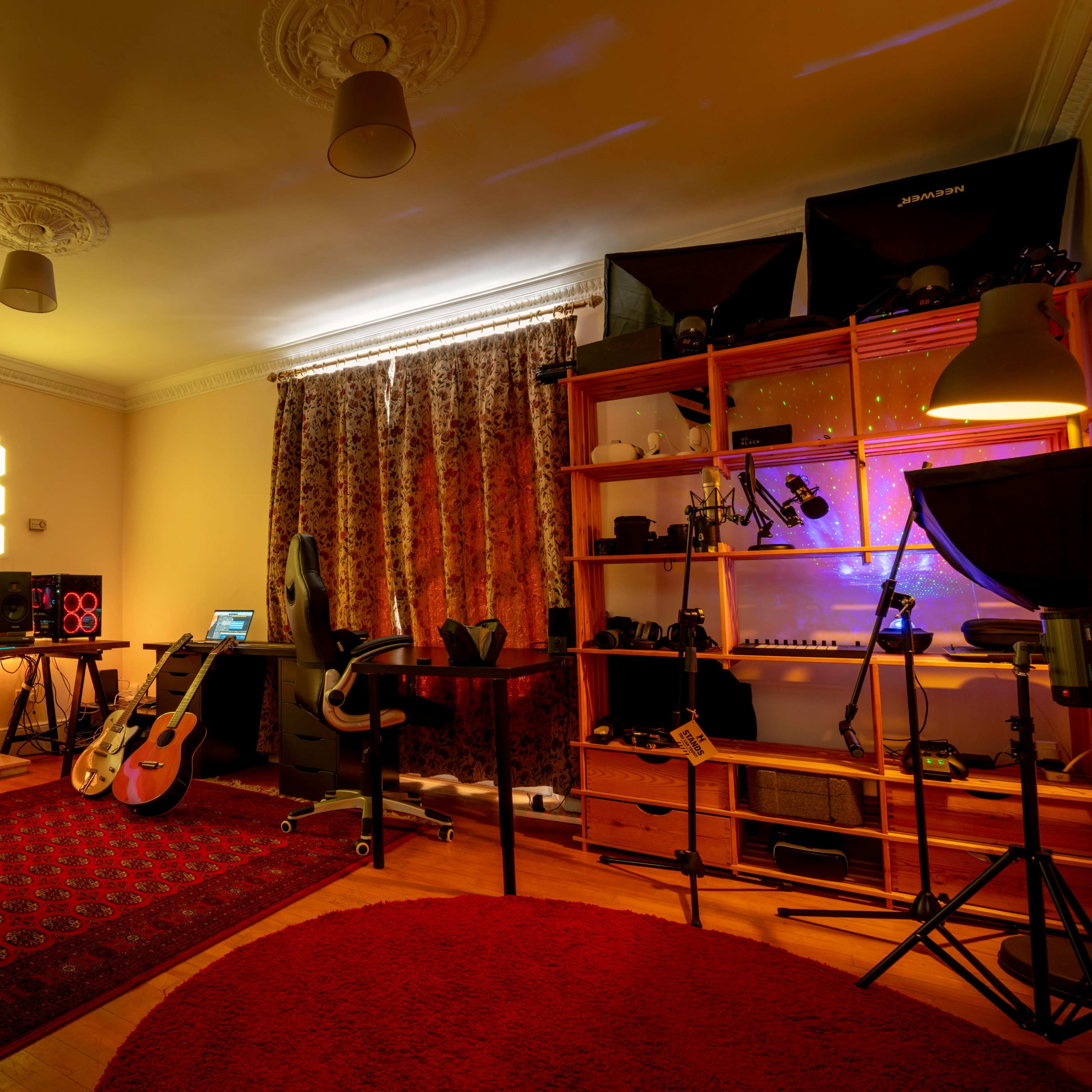 A music production studio featuring two computer monitors, musical instruments, and various equipment arranged around a cozy room with patterned curtains and a circular rug.
