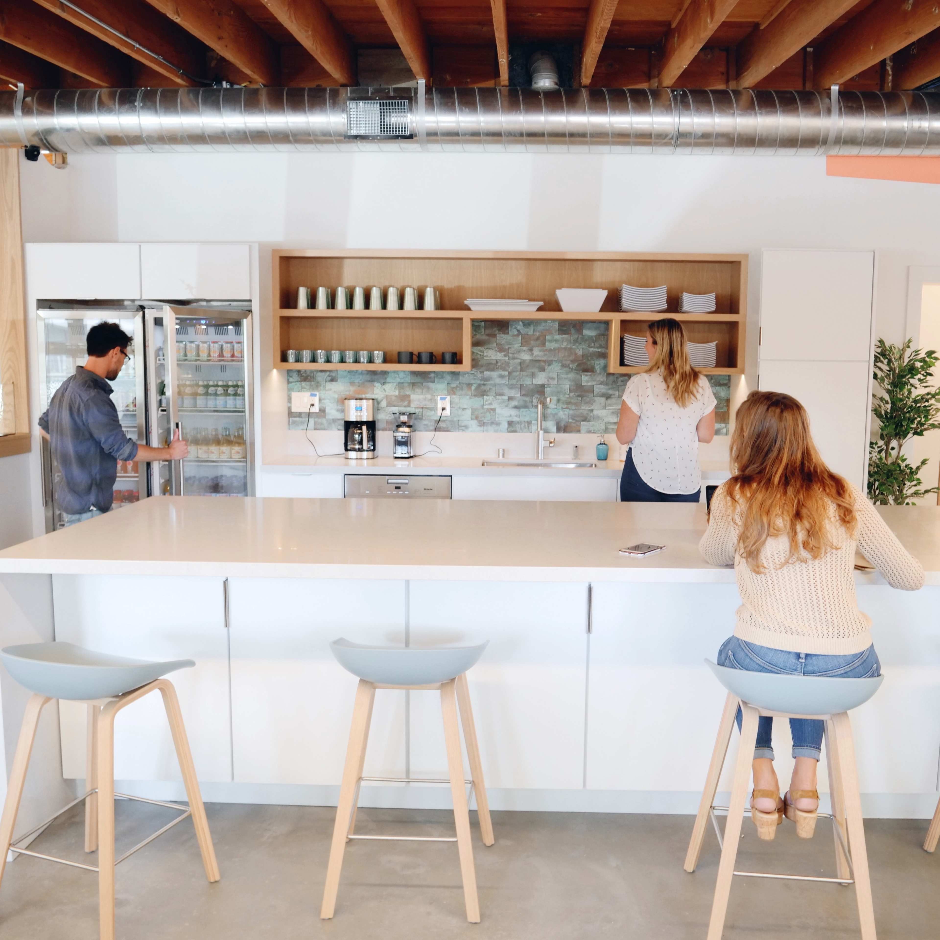 A modern kitchen features a large white island with stools, where one person sits while two others interact near a refrigerator and sink.