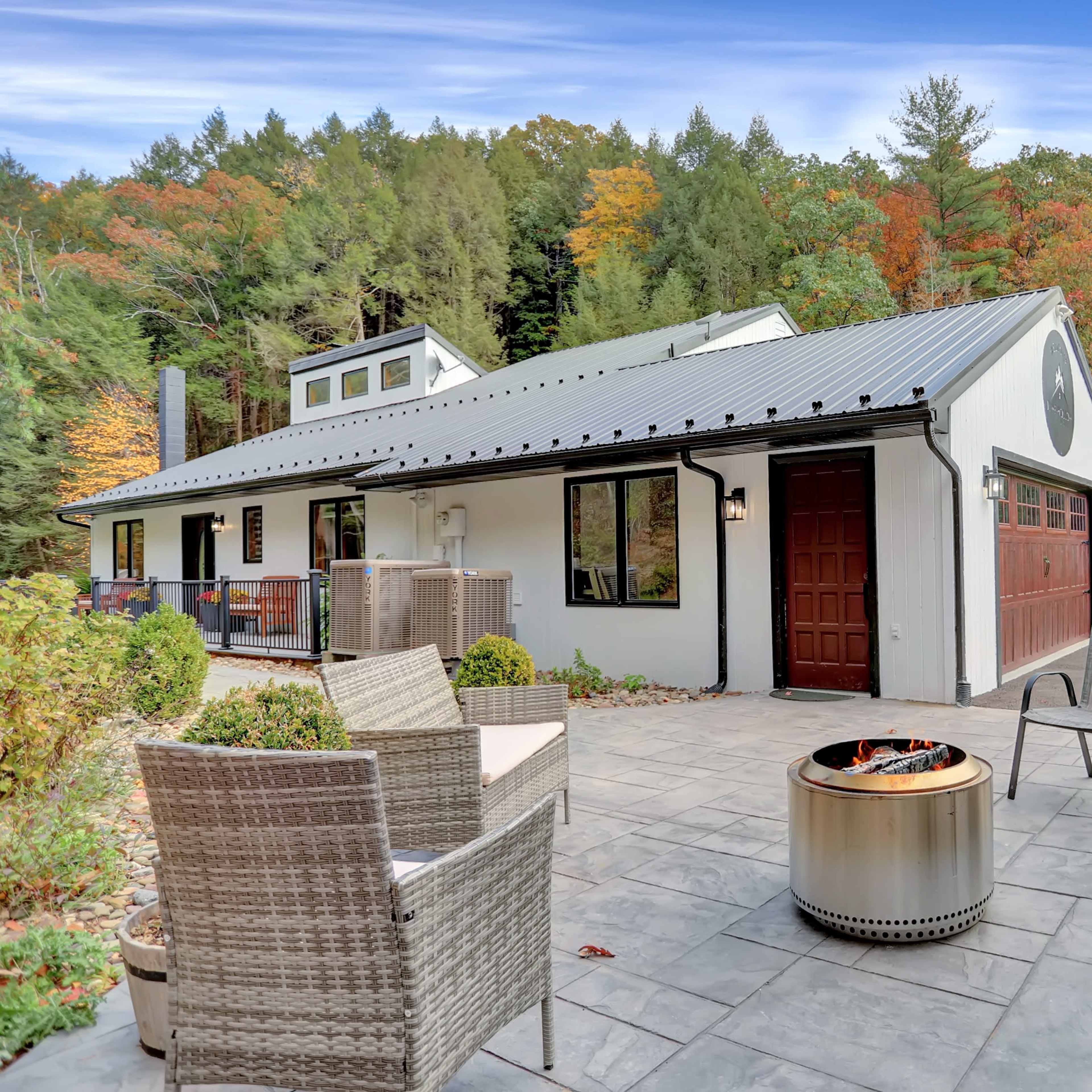 A modern house with a metal roof is situated in front of a wooded area featuring autumn foliage, and has an outdoor seating arrangement with a fire pit.