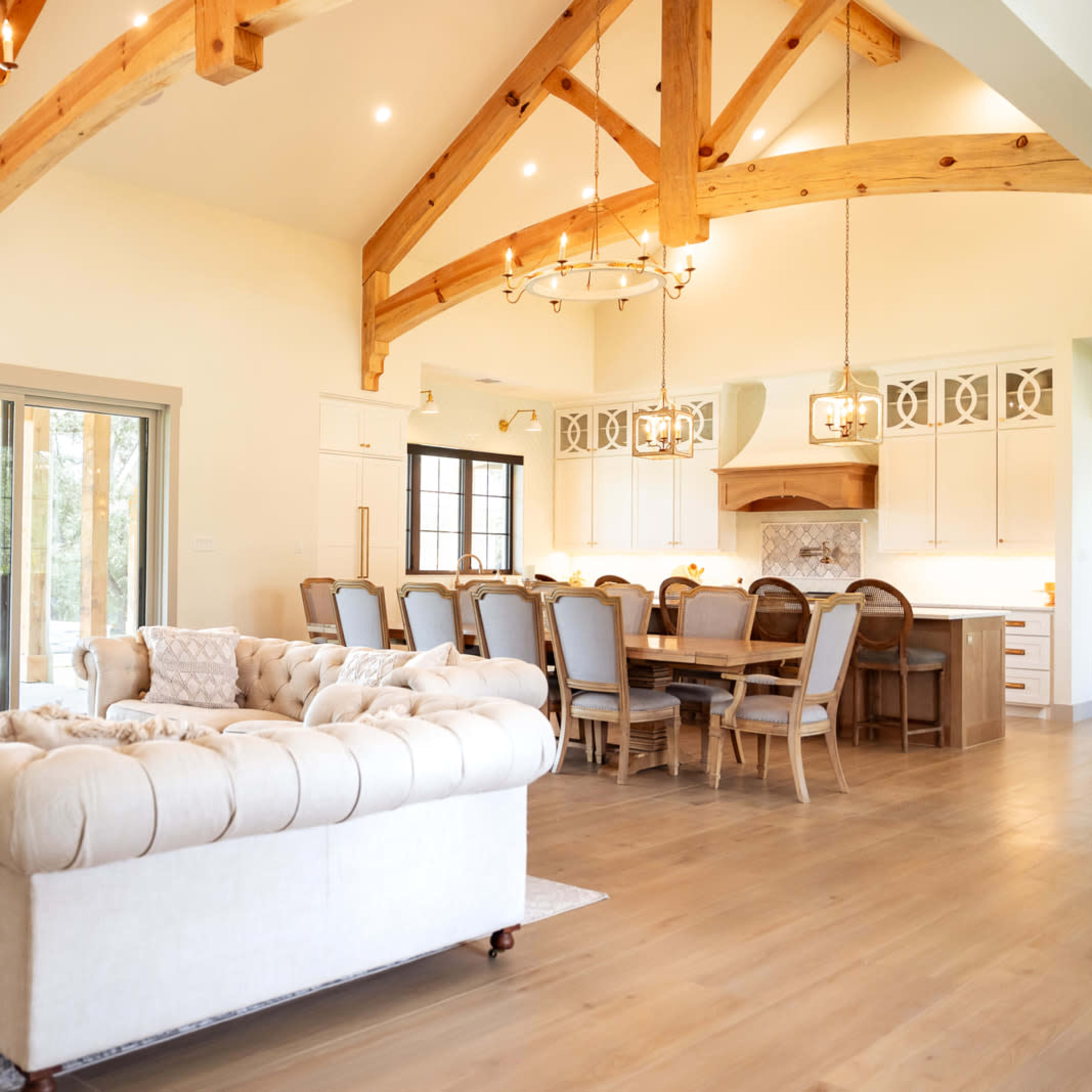 A spacious living area with a large tufted sofa, wooden beam ceilings, a dining table with chairs, and large sliding glass doors leading to an outdoor space.