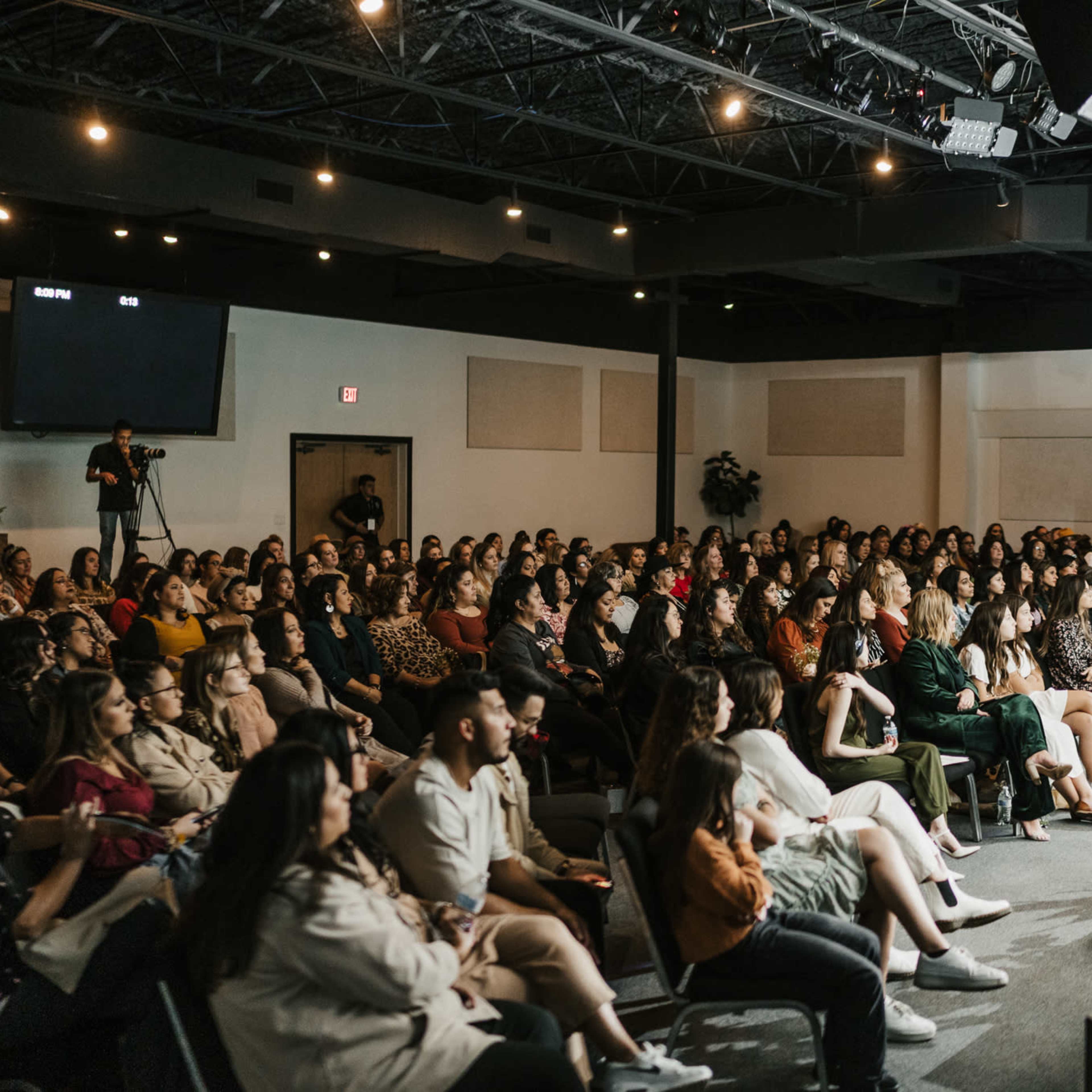 A large audience is seated in a modern lecture space, attentively facing a stage with a speaker and a screen.