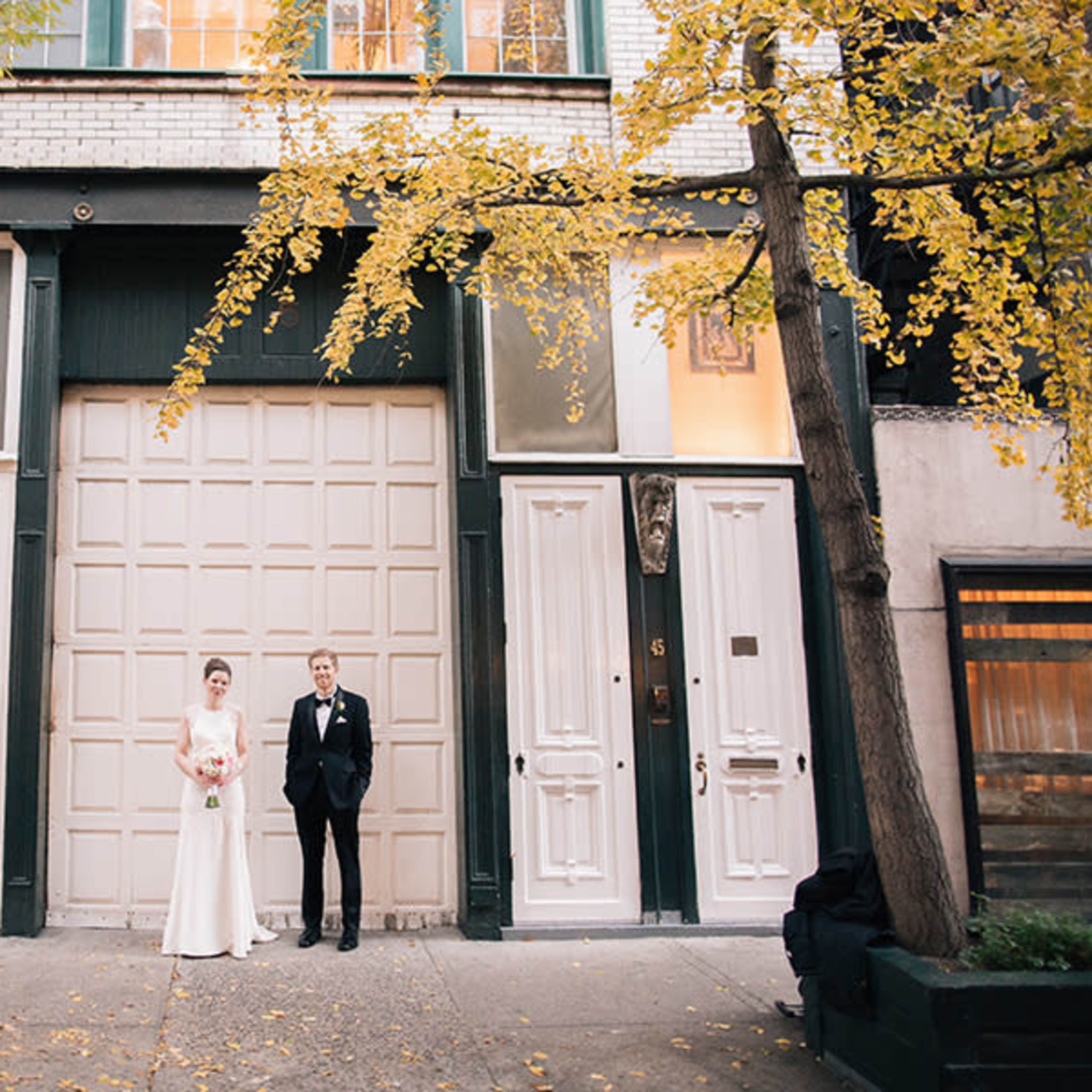A bride and groom stand in front of a building with a light-colored door and tall windows, framed by autumn trees with yellow leaves.