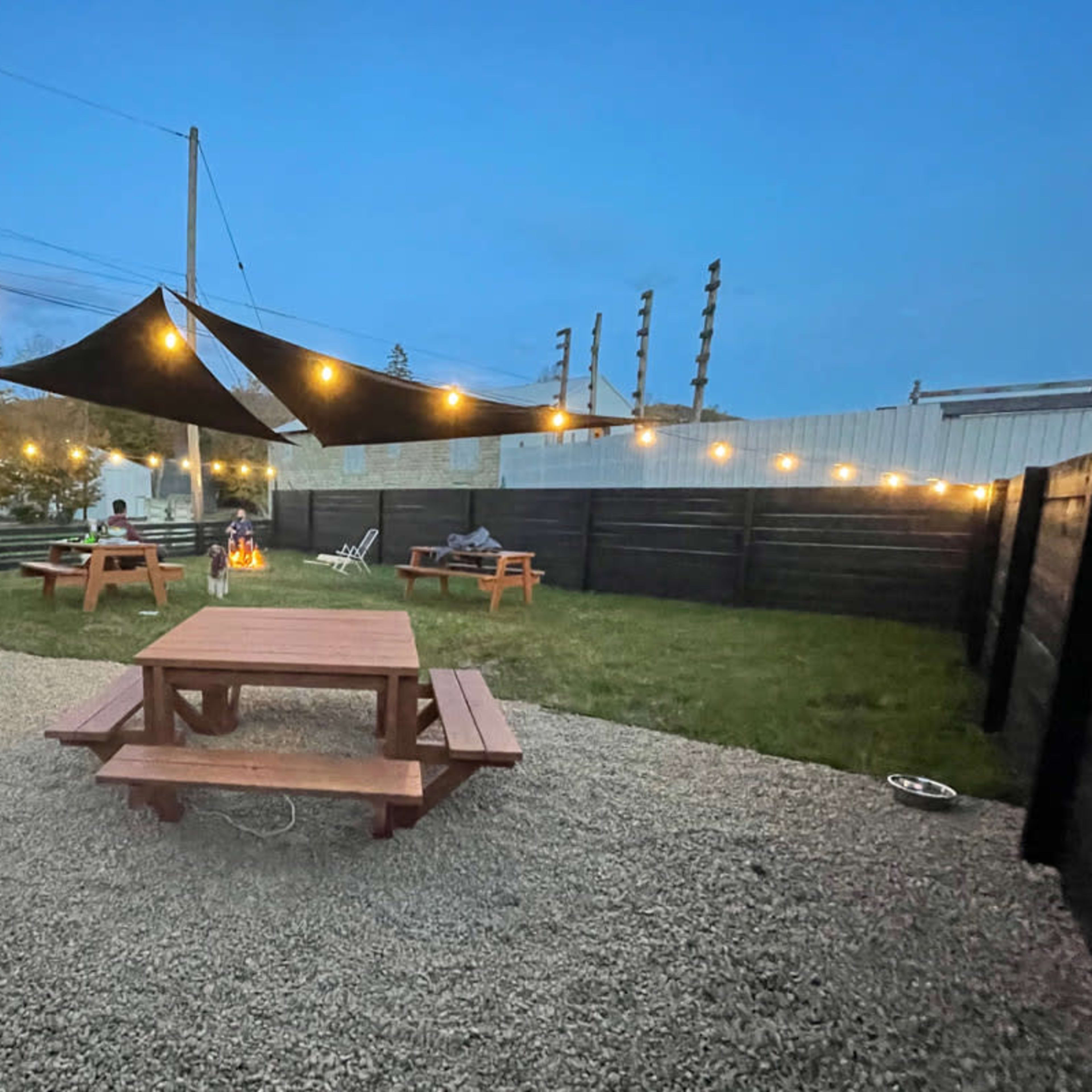 A gravel outdoor space with wooden picnic tables, string lights, and a fire pit surrounded by black wooden fences.