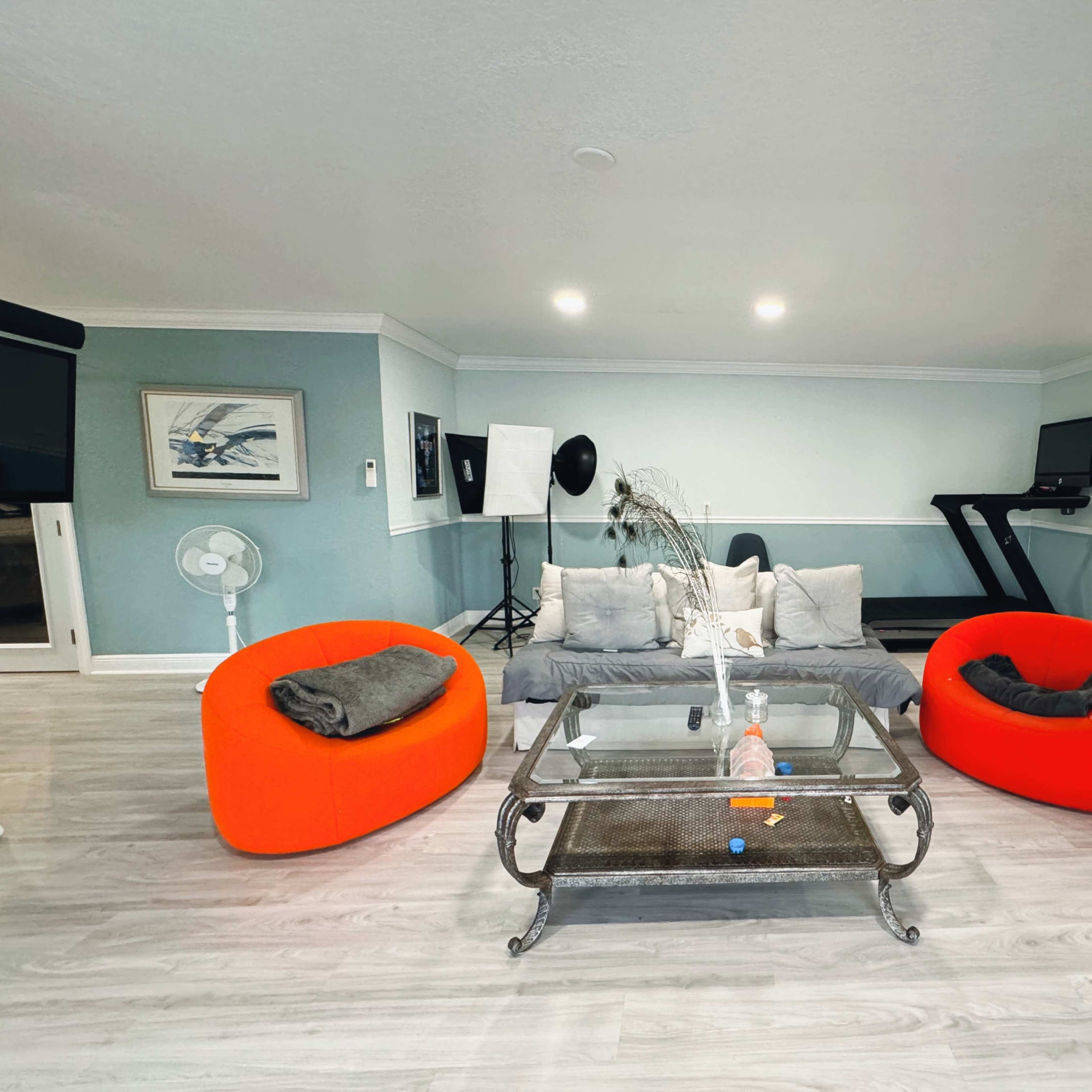 A modern living area features two bright red chairs, a glass coffee table, and a treadmill, with light-colored walls and a fan in the background.
