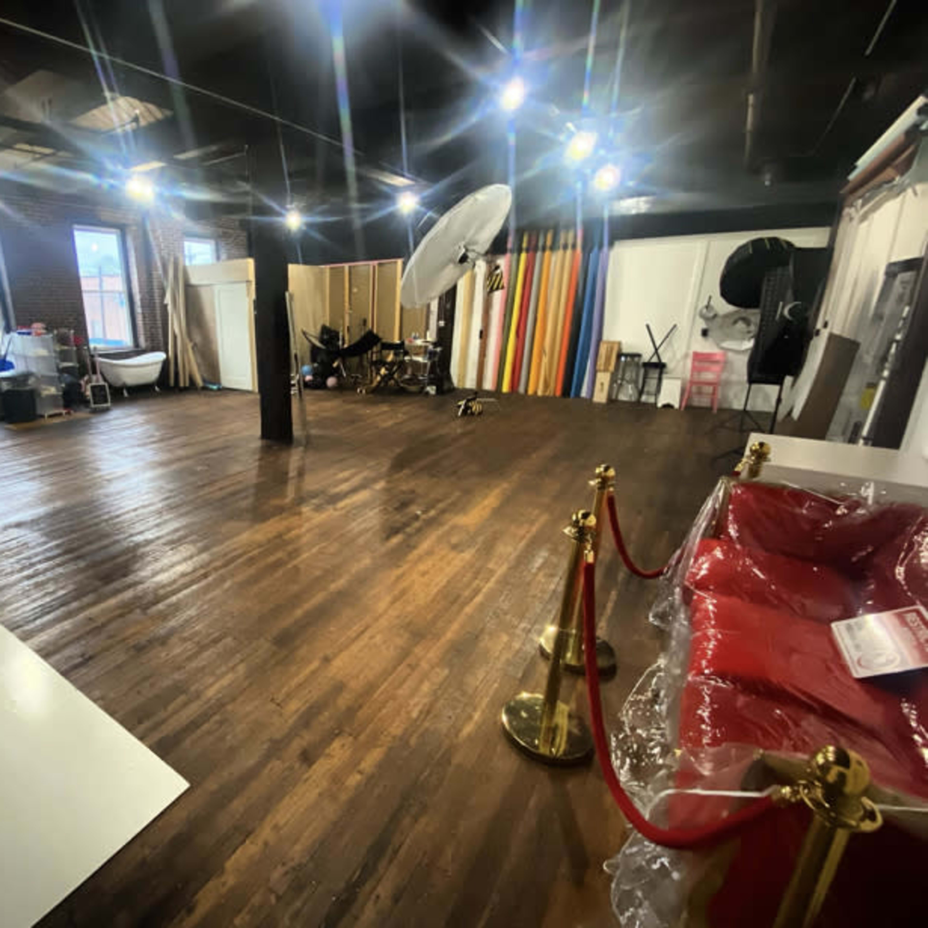 A spacious studio with wooden floors, various backdrops in the background, and a red sofa with a velvet rope in the foreground.