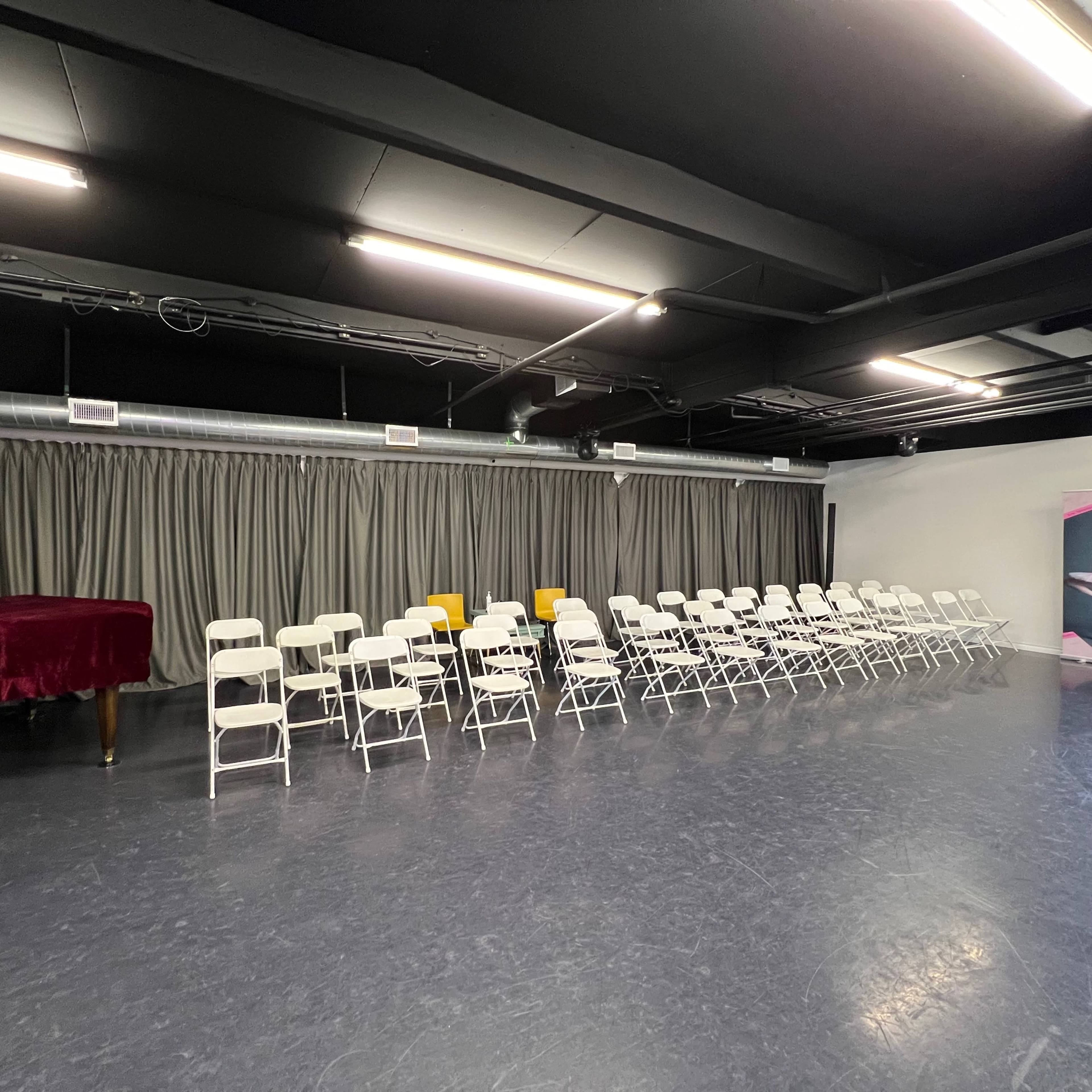 A dance studio with rows of white chairs, a red piano, and a curtain along one wall.