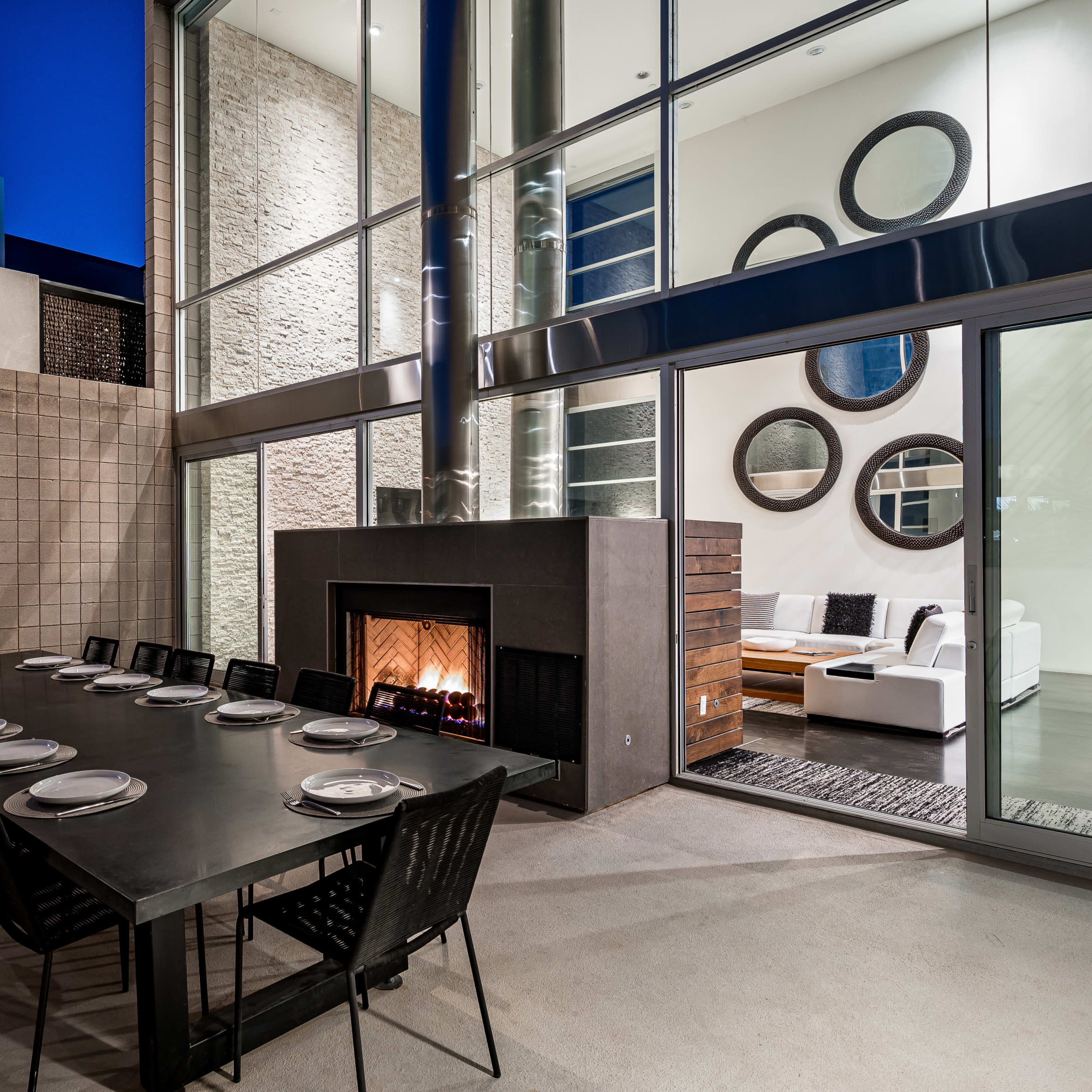 A modern outdoor dining area features a long table set for dinner next to a glass-enclosed living space with a fireplace and circular mirrors.