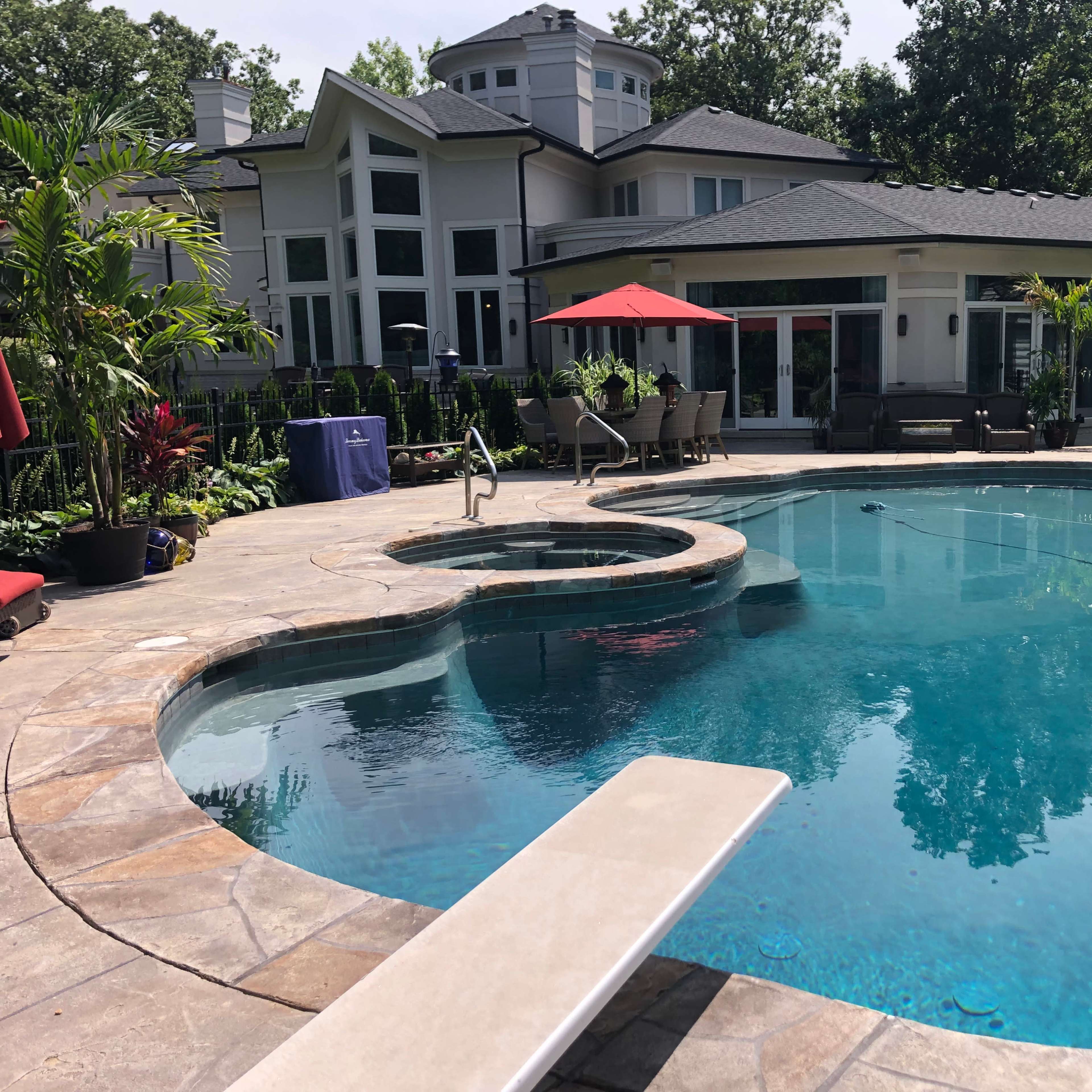 A residential backyard with a swimming pool, a diving board, and a waterslide beside a large house surrounded by greenery.