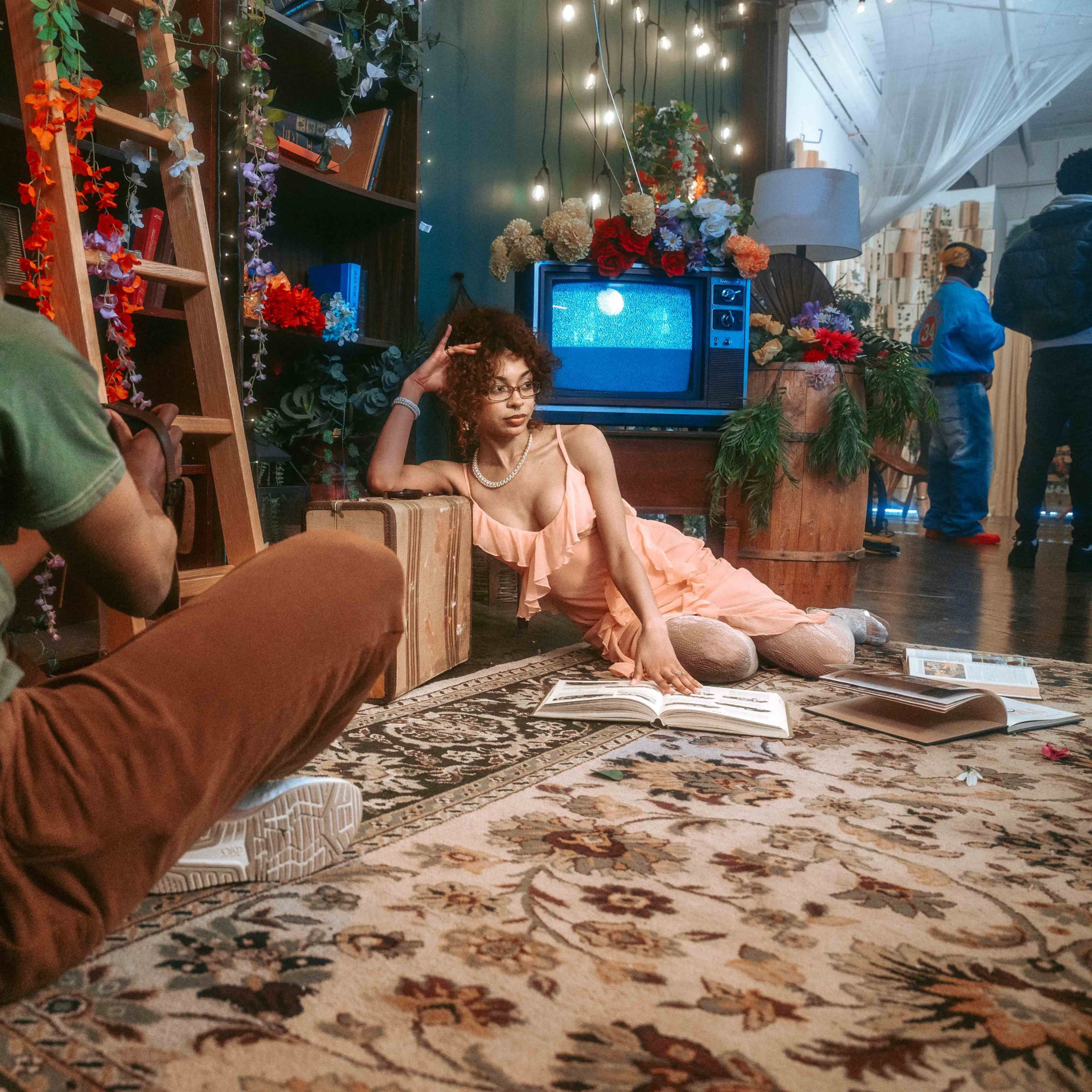 A photographer captures a model posing on a carpeted floor surrounded by floral decorations and bookshelves in a well-lit indoor setting.