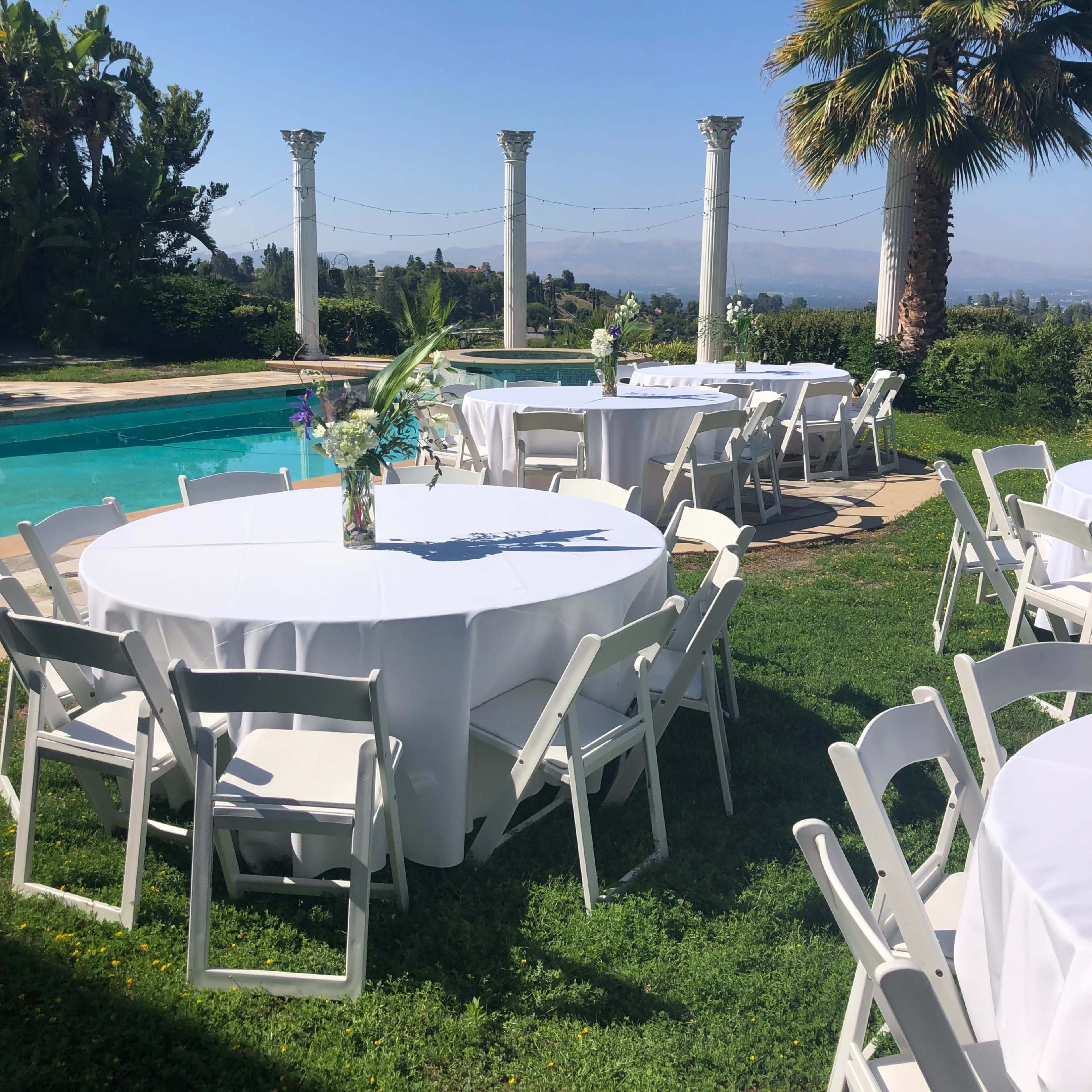 A garden setting for an outdoor event, featuring round tables with white tablecloths arranged near a swimming pool and ornamental columns in the background.