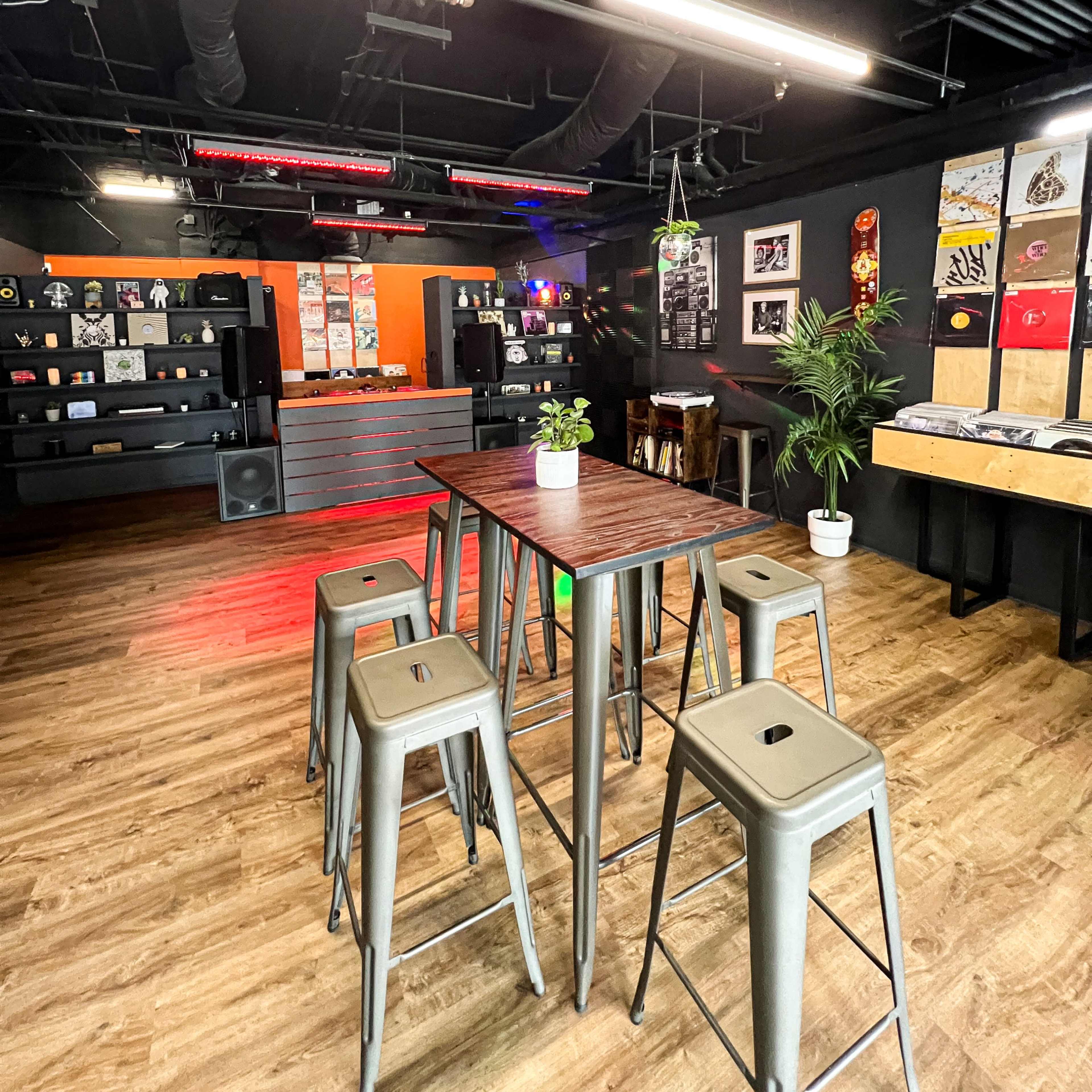 A modern lounge area with a high table surrounded by metal stools, wooden flooring, a wall displaying various artworks, and audio equipment in the background.