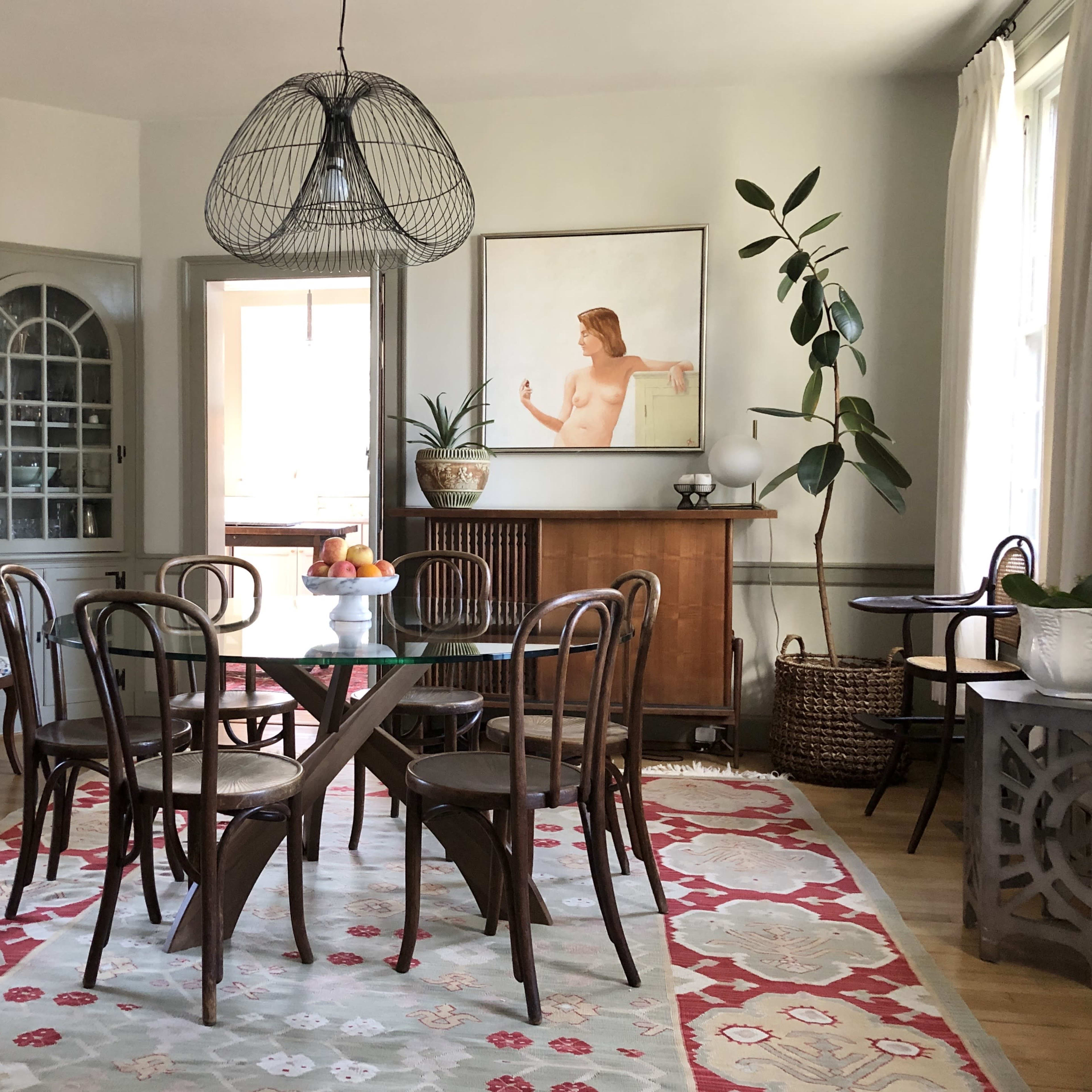 A dining area features a round table with chairs, a large artistic wall piece, and plants beside a sideboard in a well-lit room.