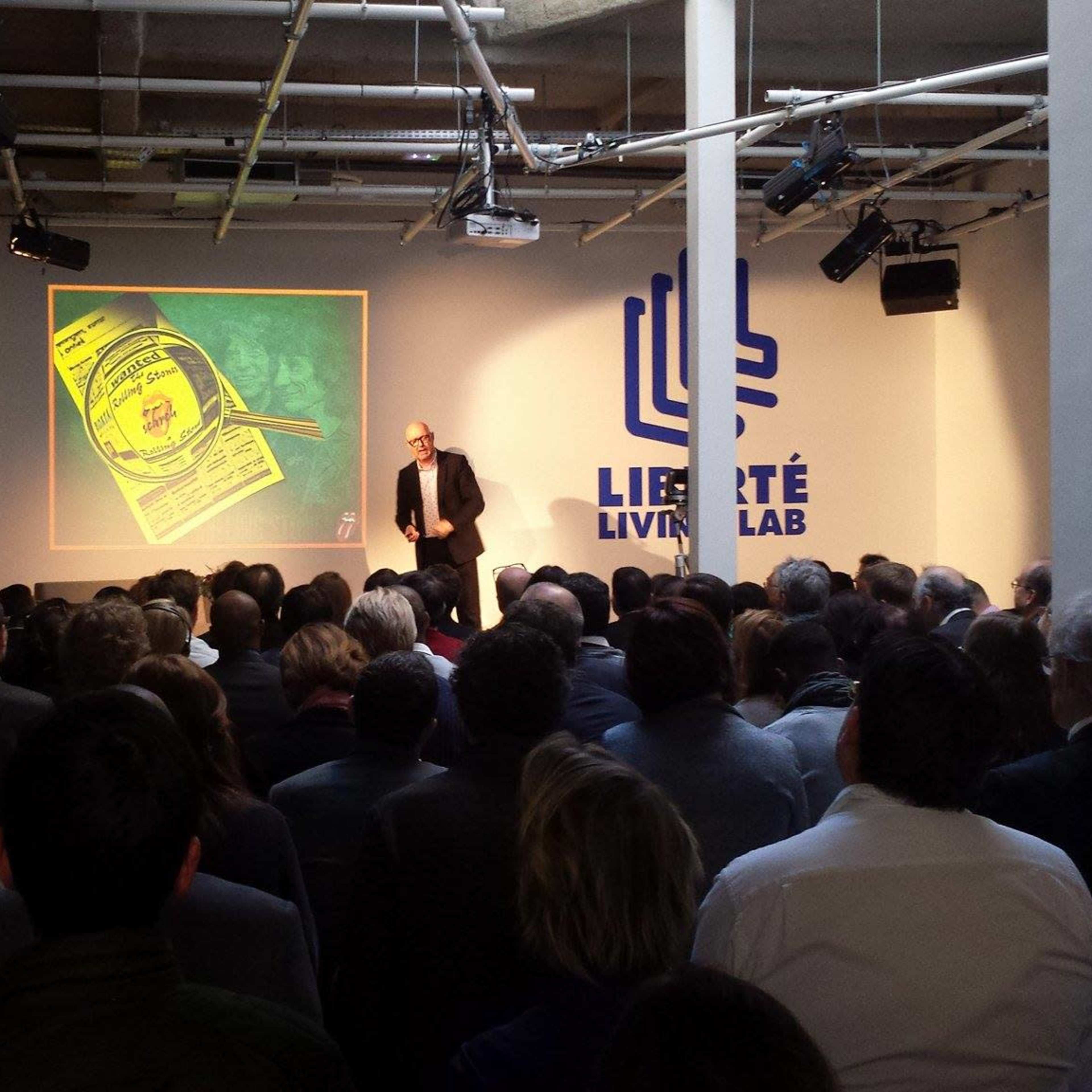A speaker presents in front of an audience at a venue labeled "Liberté Living Lab," with a visual aid projected on the screen behind them.