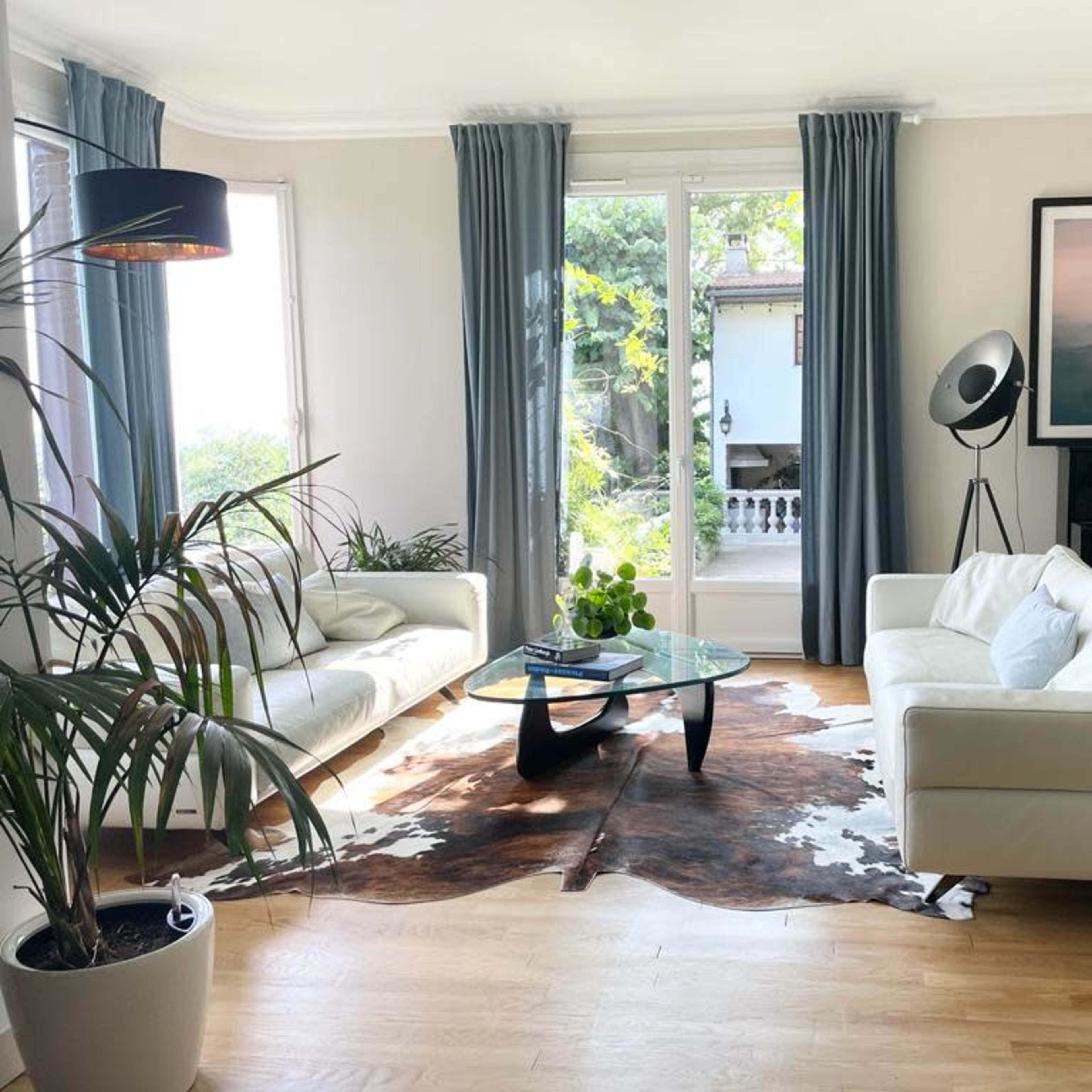 A bright living room features two white sofas facing a glass coffee table, with a large window, potted plants, and a modern art piece on the wall.