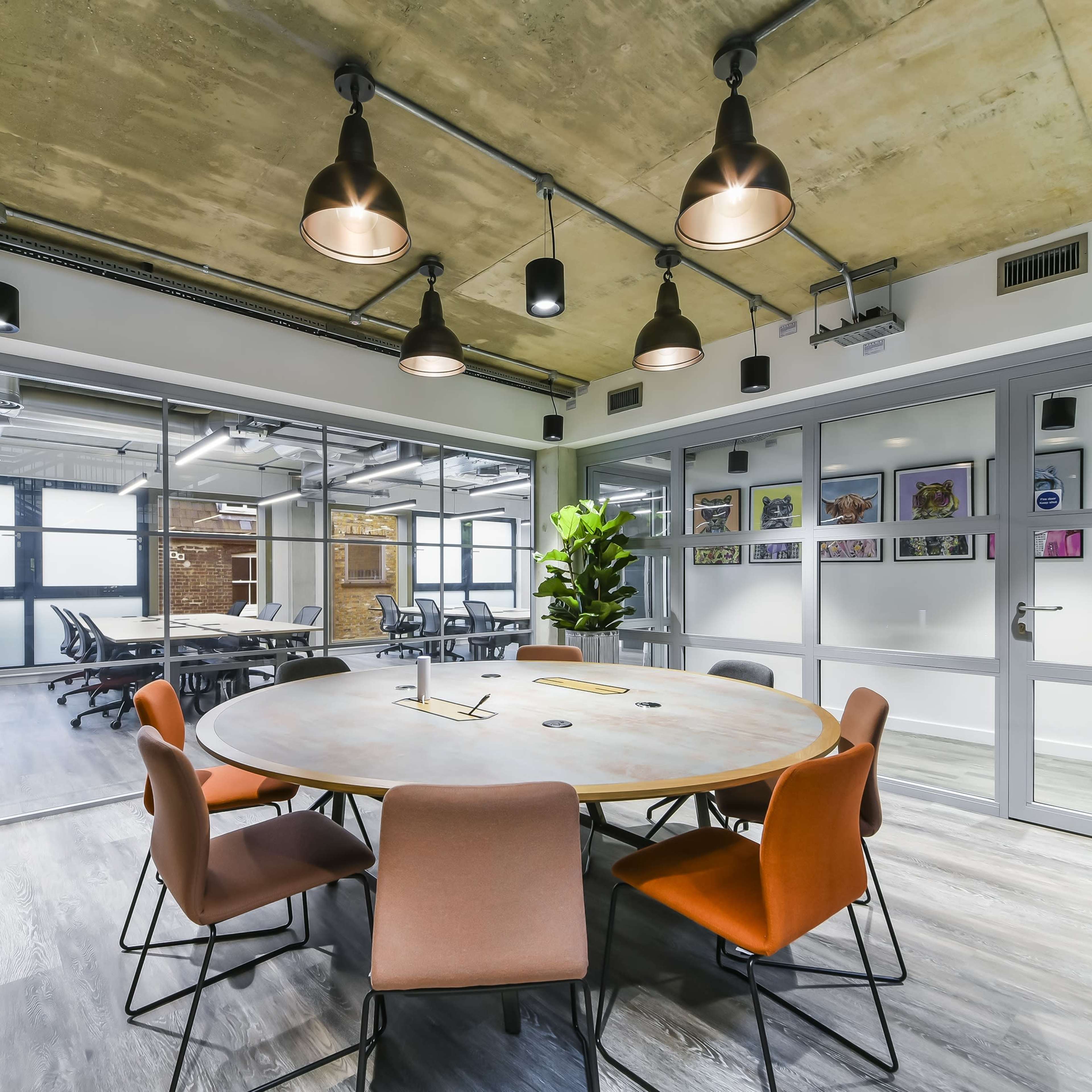 A round table surrounded by eight chairs is placed in a modern office space with glass walls and overhead lighting.
