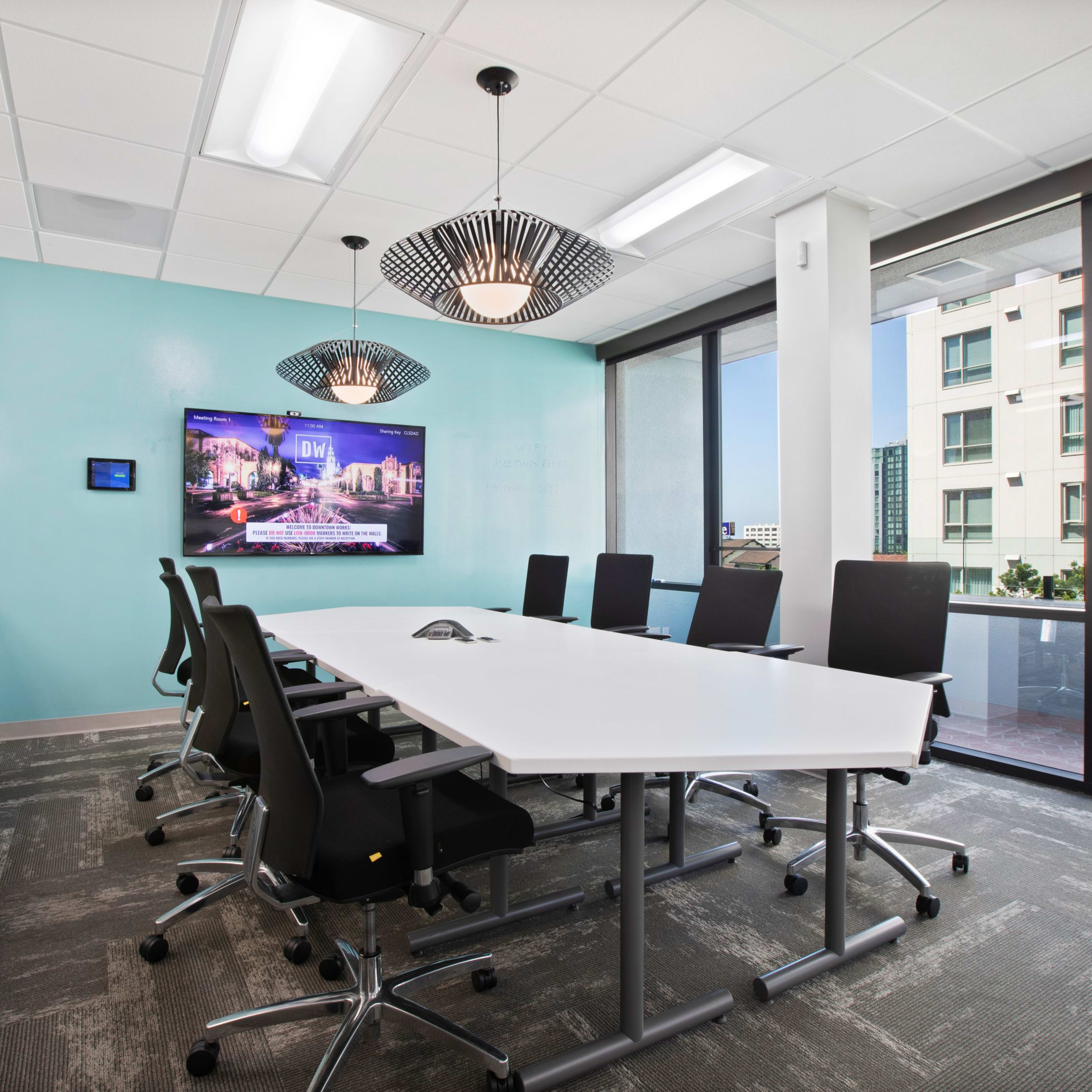 A modern conference room features a large table surrounded by black chairs, a wall-mounted screen displaying a cityscape, and large windows overlooking an outdoor area.