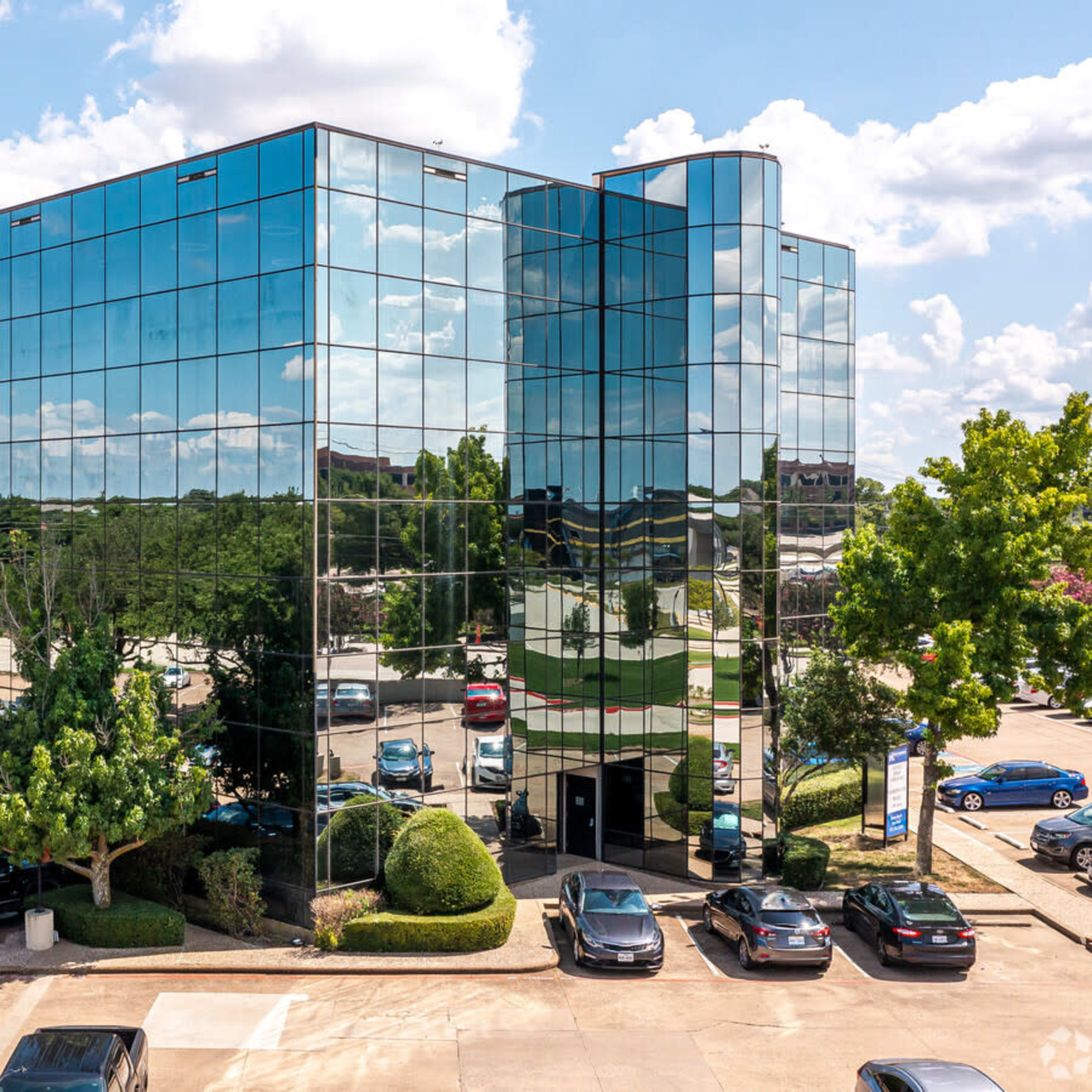 A modern glass office building with reflective windows stands among parked cars and trees under a blue sky with clouds.