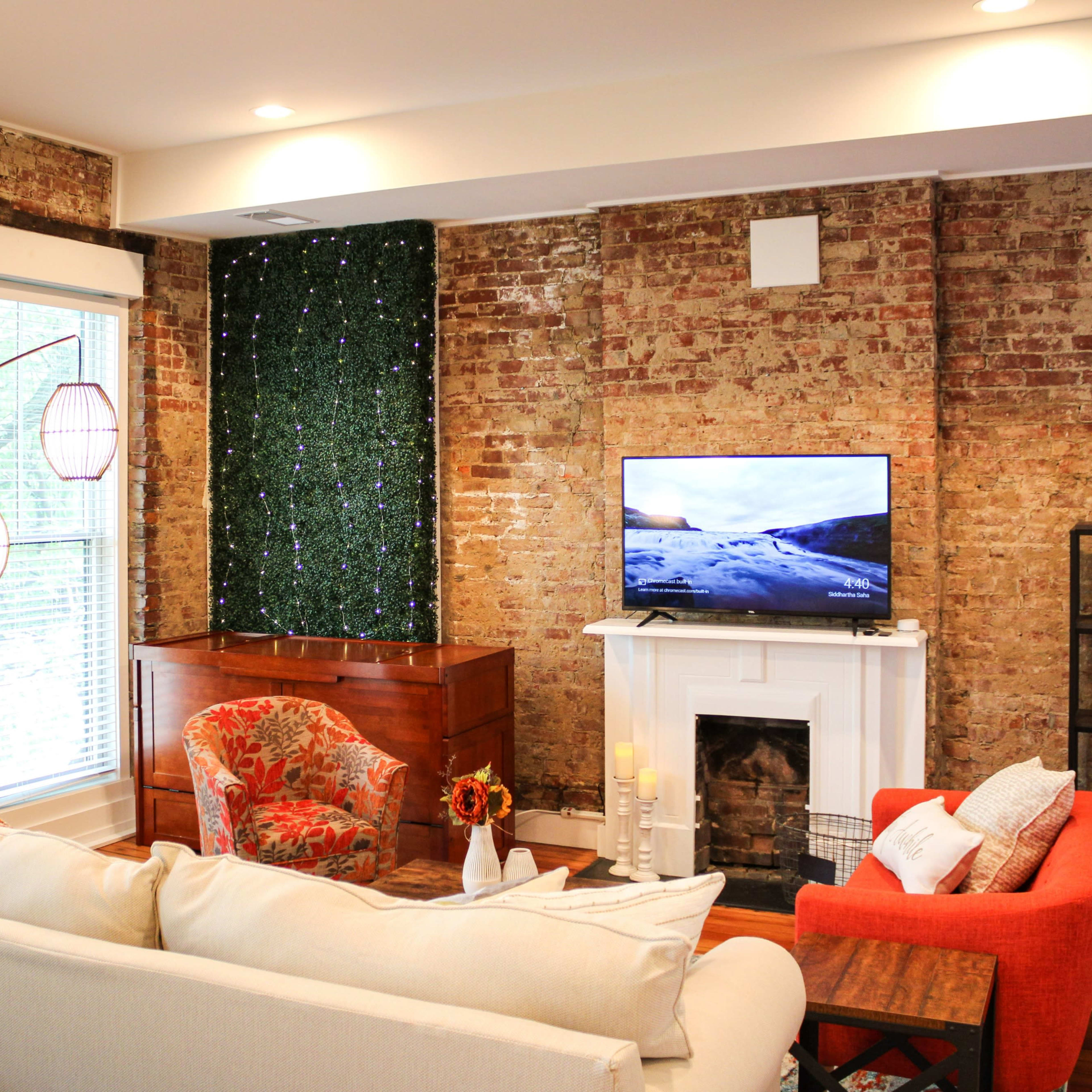 A cozy living room features exposed brick walls, a television on a stand, and a sectional sofa opposite a wooden chest and a red armchair.