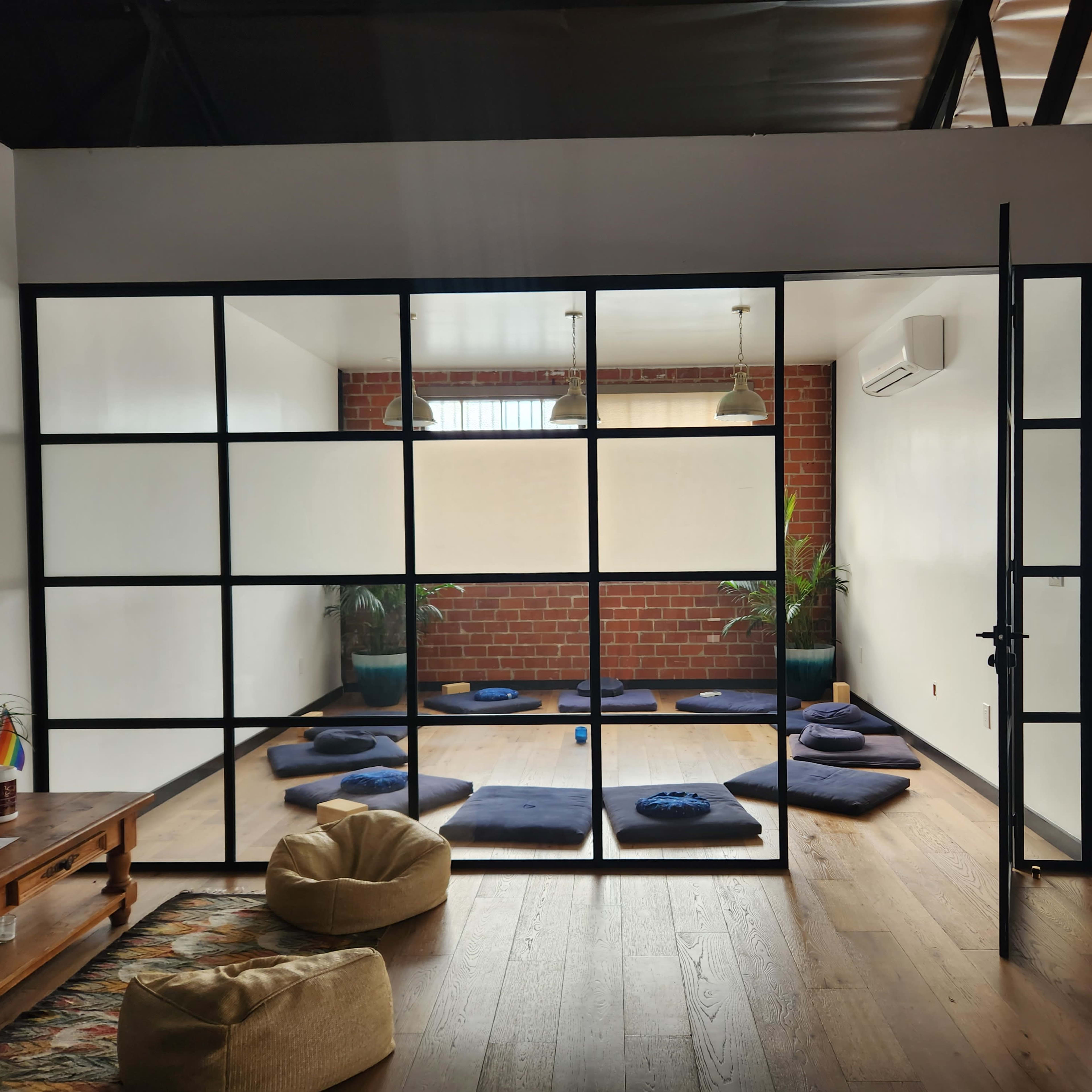 A spacious room with wooden flooring features meditation mats arranged on the floor, framed by a glass wall and greenery from potted plants.