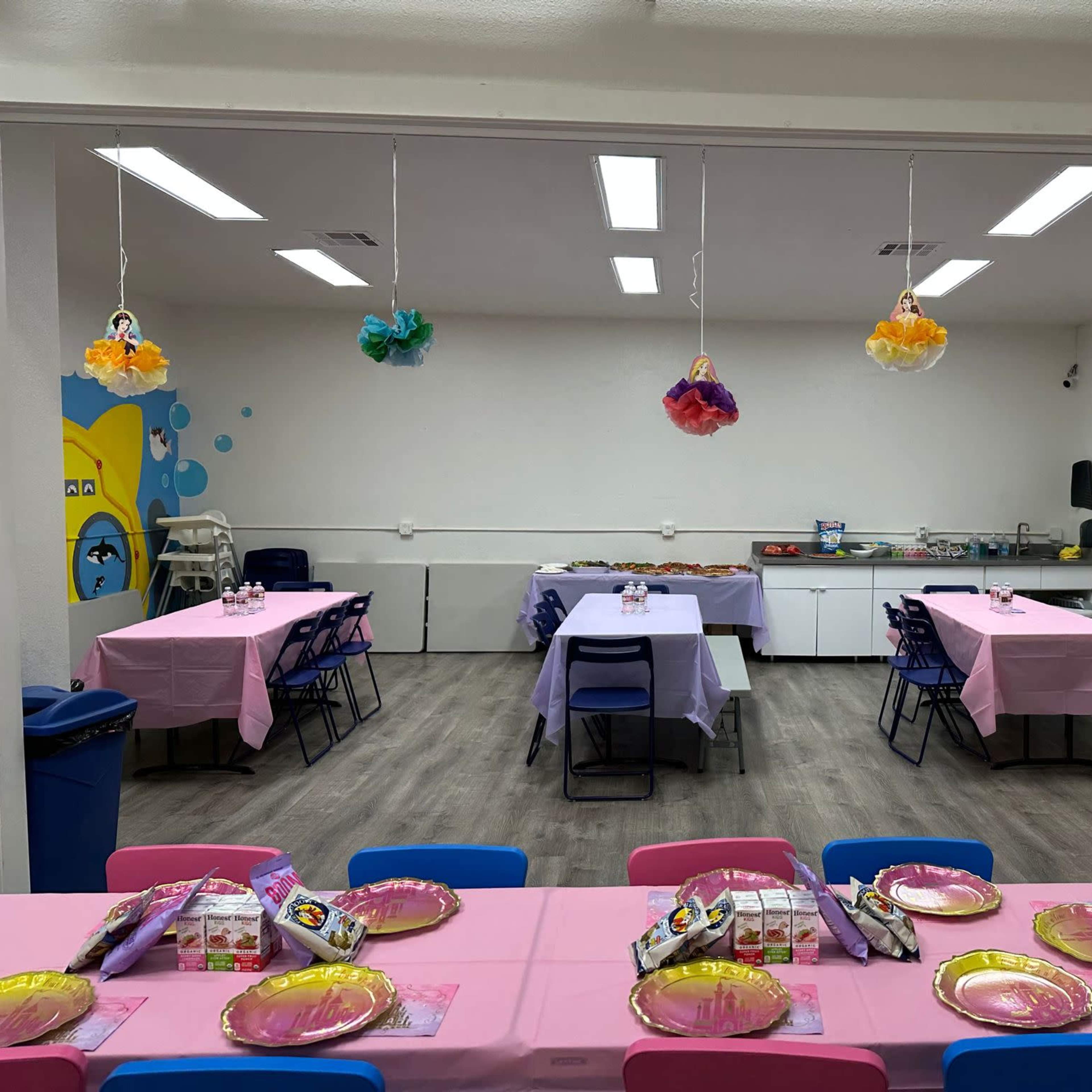 A brightly lit room set up for a party, featuring several tables covered with pink tablecloths and decorated with colorful hanging decorations.