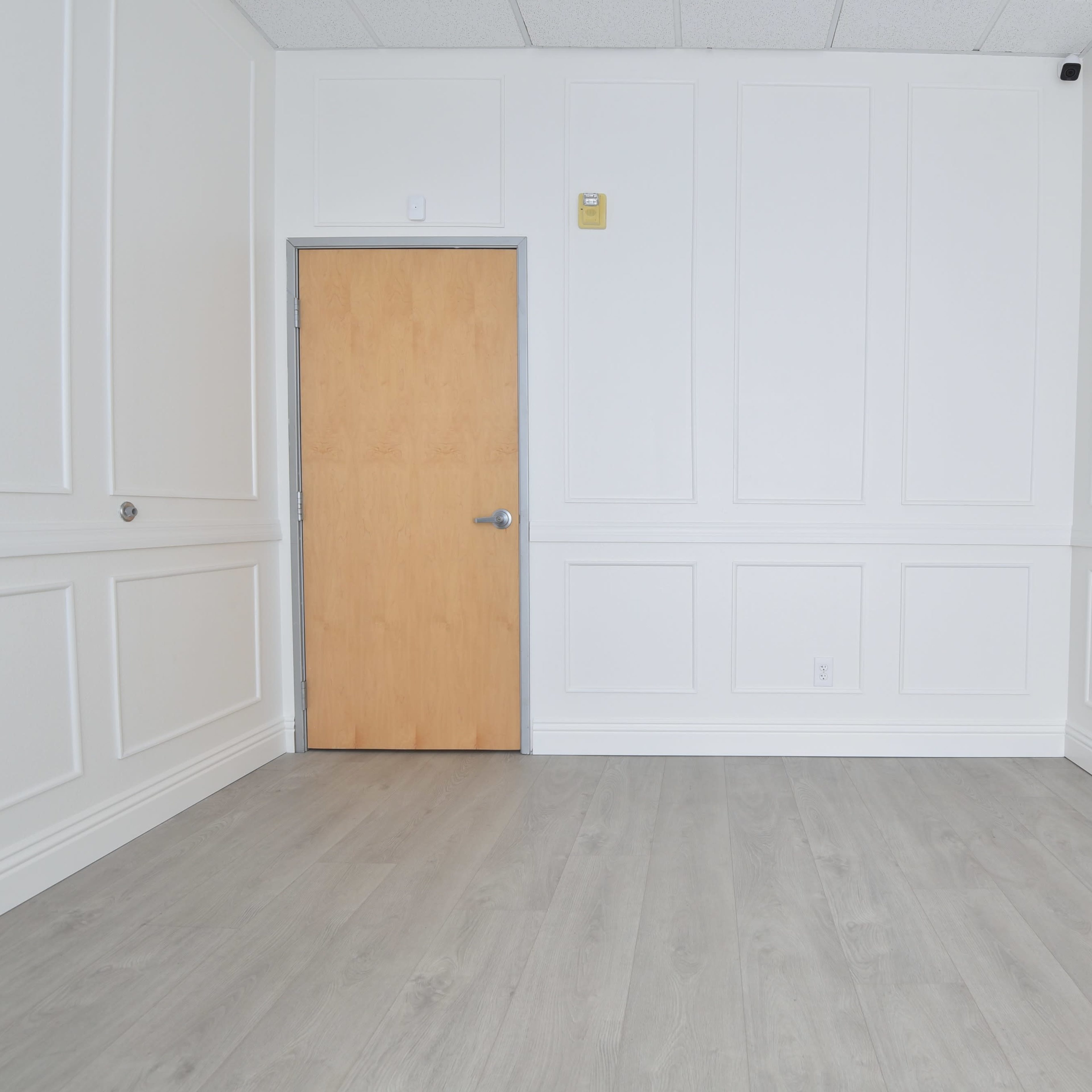 A sparse room with a light-colored wooden door at one end and plain white walls with paneling.
