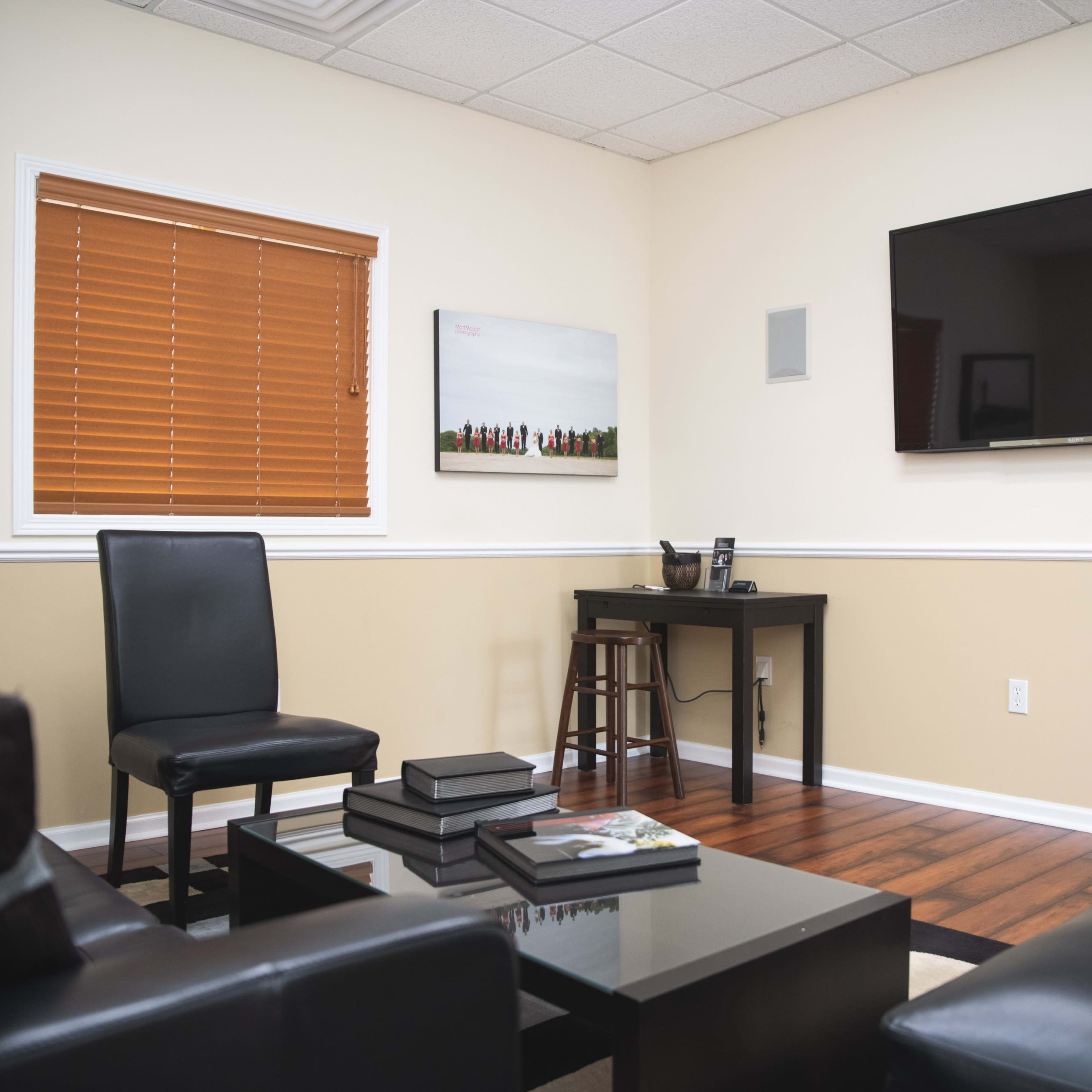 A small, modern office or meeting room with a television mounted on the wall, a black desk, a bar stool, and a few black chairs arranged around a coffee table.