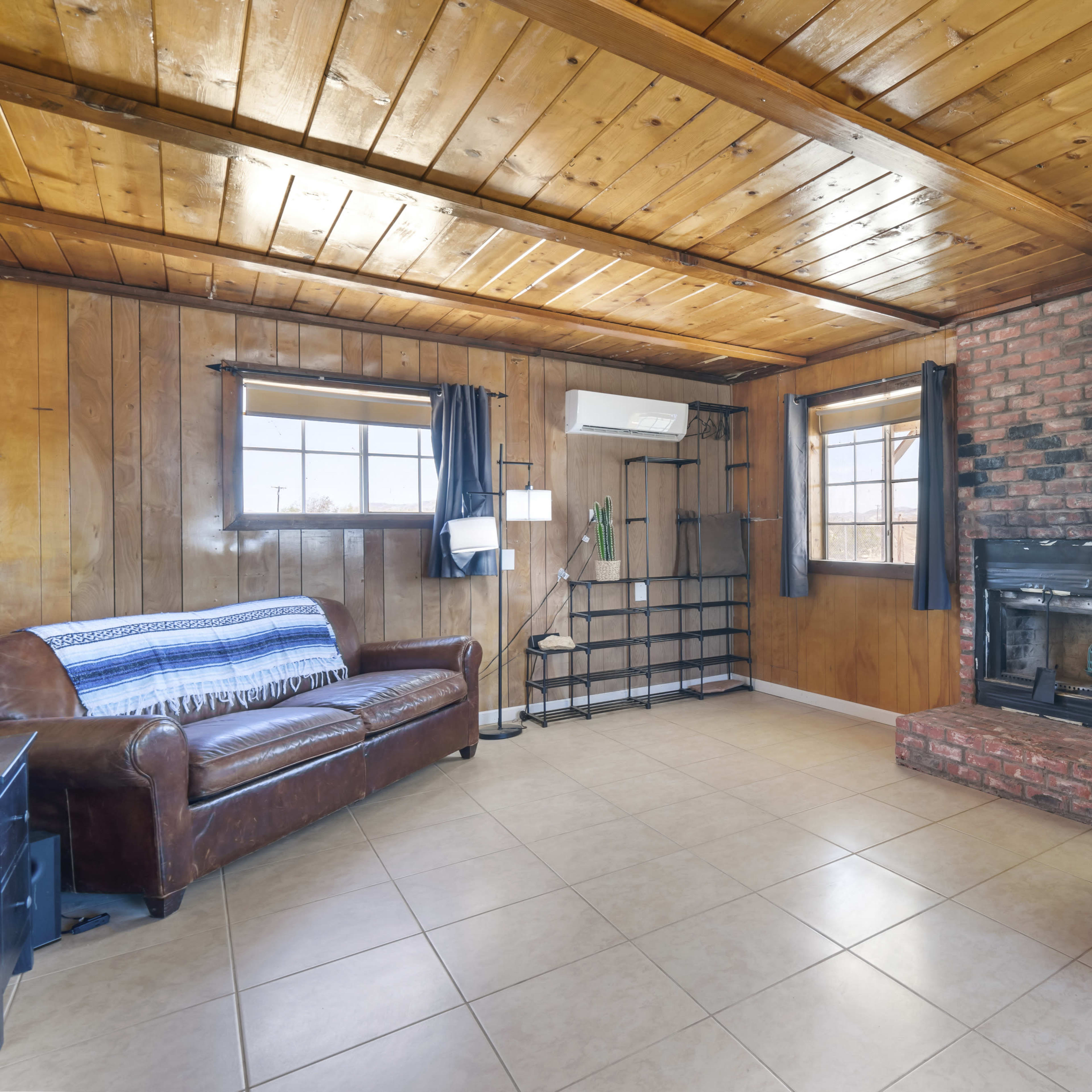 The living room features wooden walls and a ceiling, a brick fireplace, a leather sofa, and a blue upholstered chair, with large windows allowing natural light.