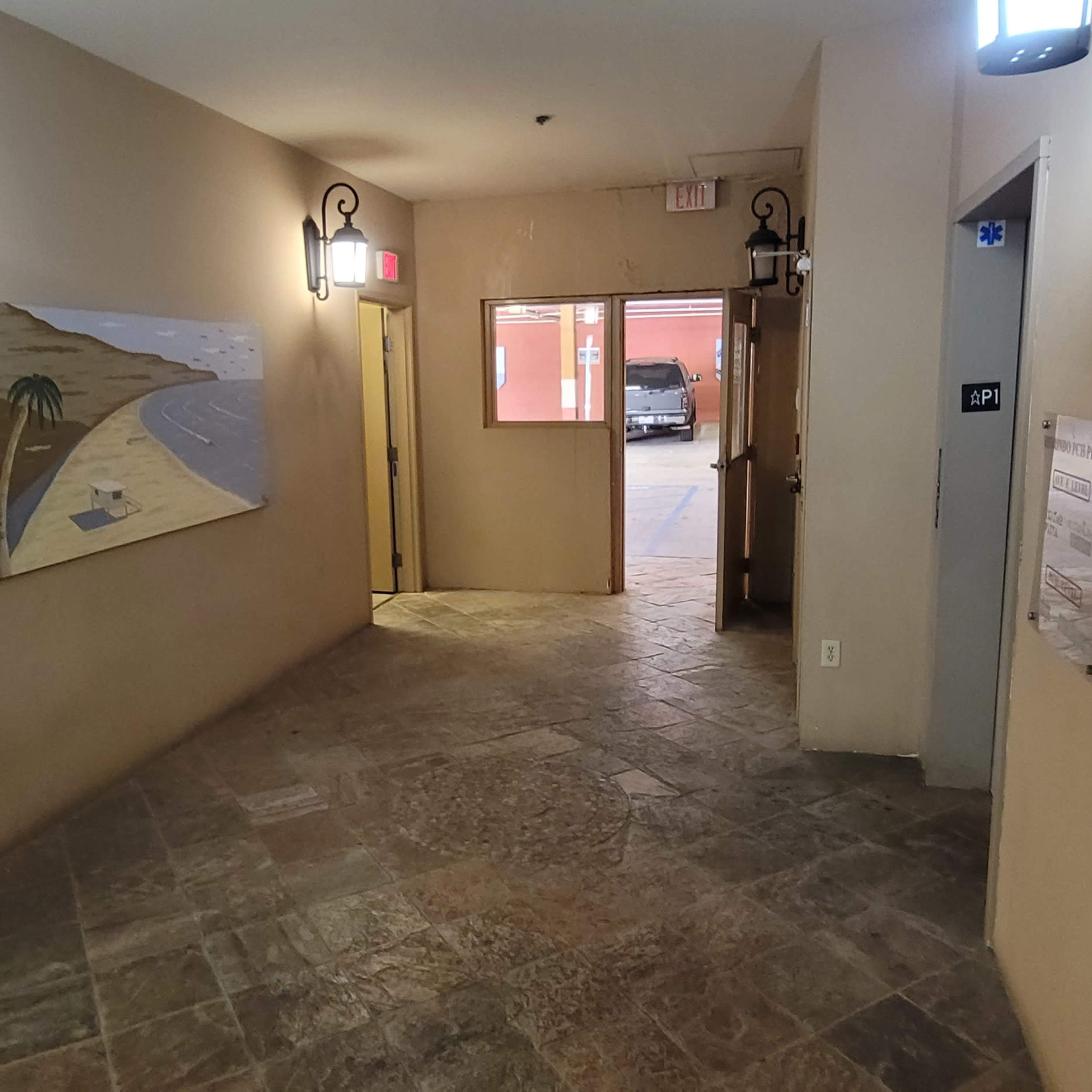 A hallway with tiled flooring, a painting on the wall, and doors leading to an outdoor area.
