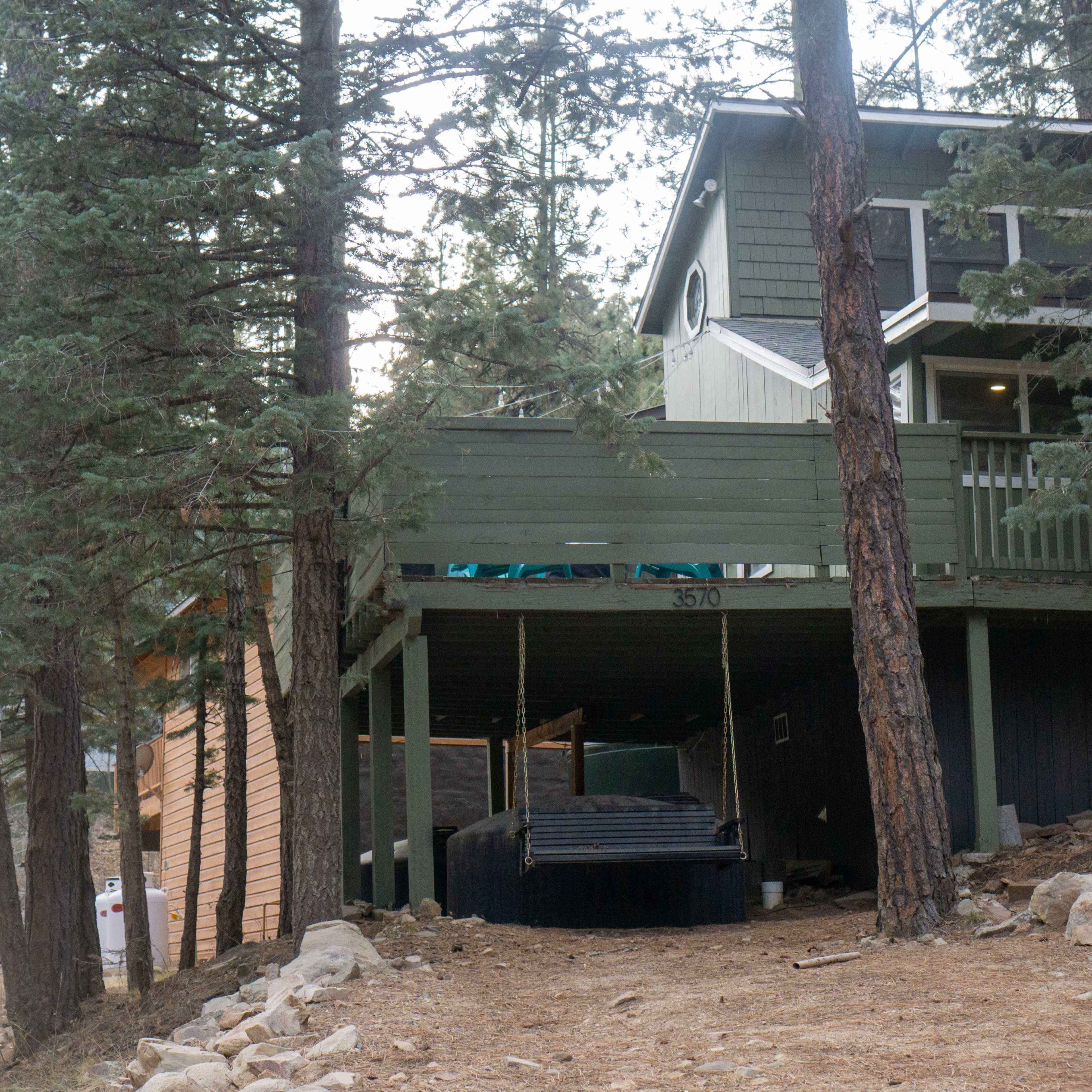 A house with a wraparound porch is situated among pine trees, with a swing hanging underneath the elevated structure.