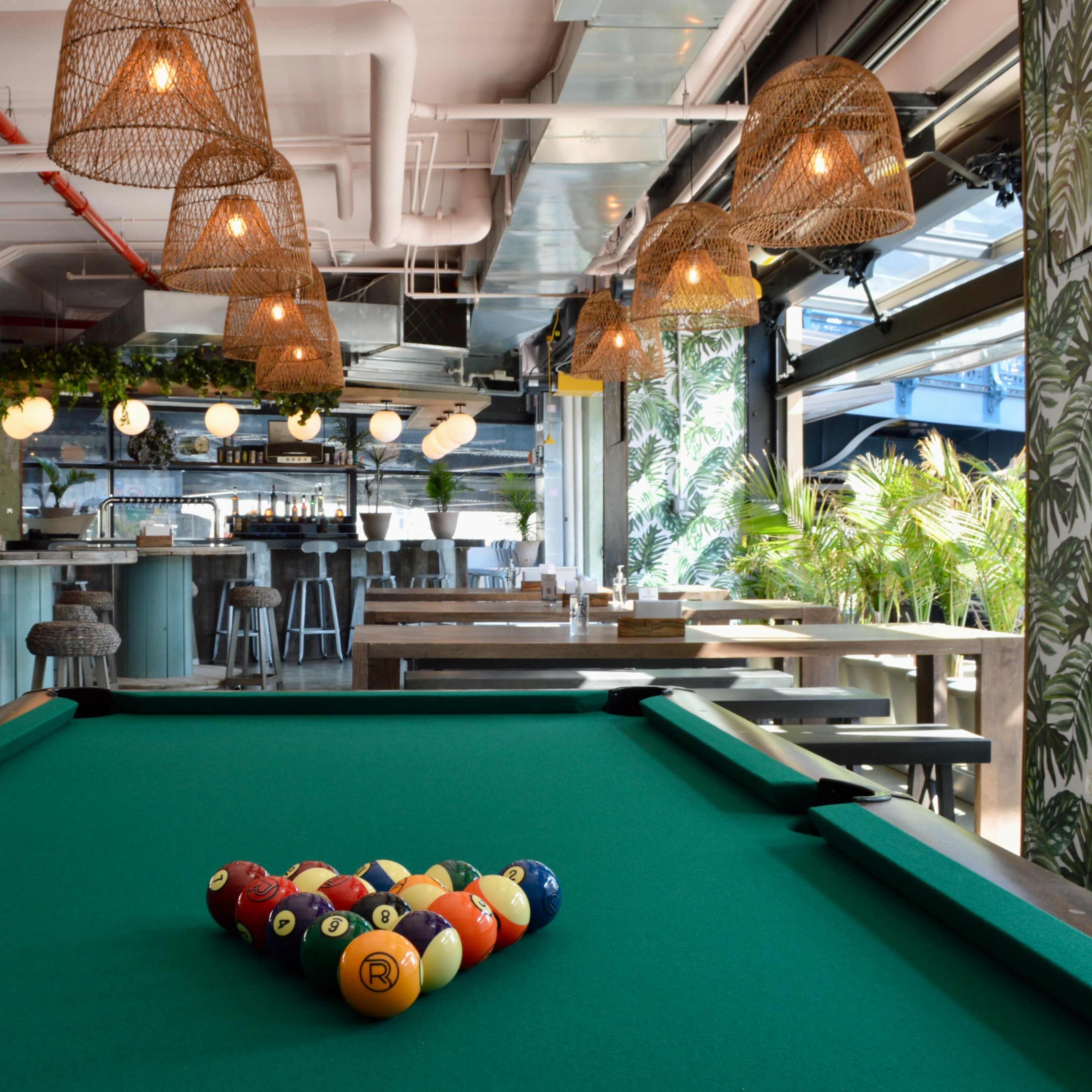 A pool table with a neatly arranged set of colored balls occupies the foreground of a modern, well-lit bar and dining space featuring hanging light fixtures and green plants.