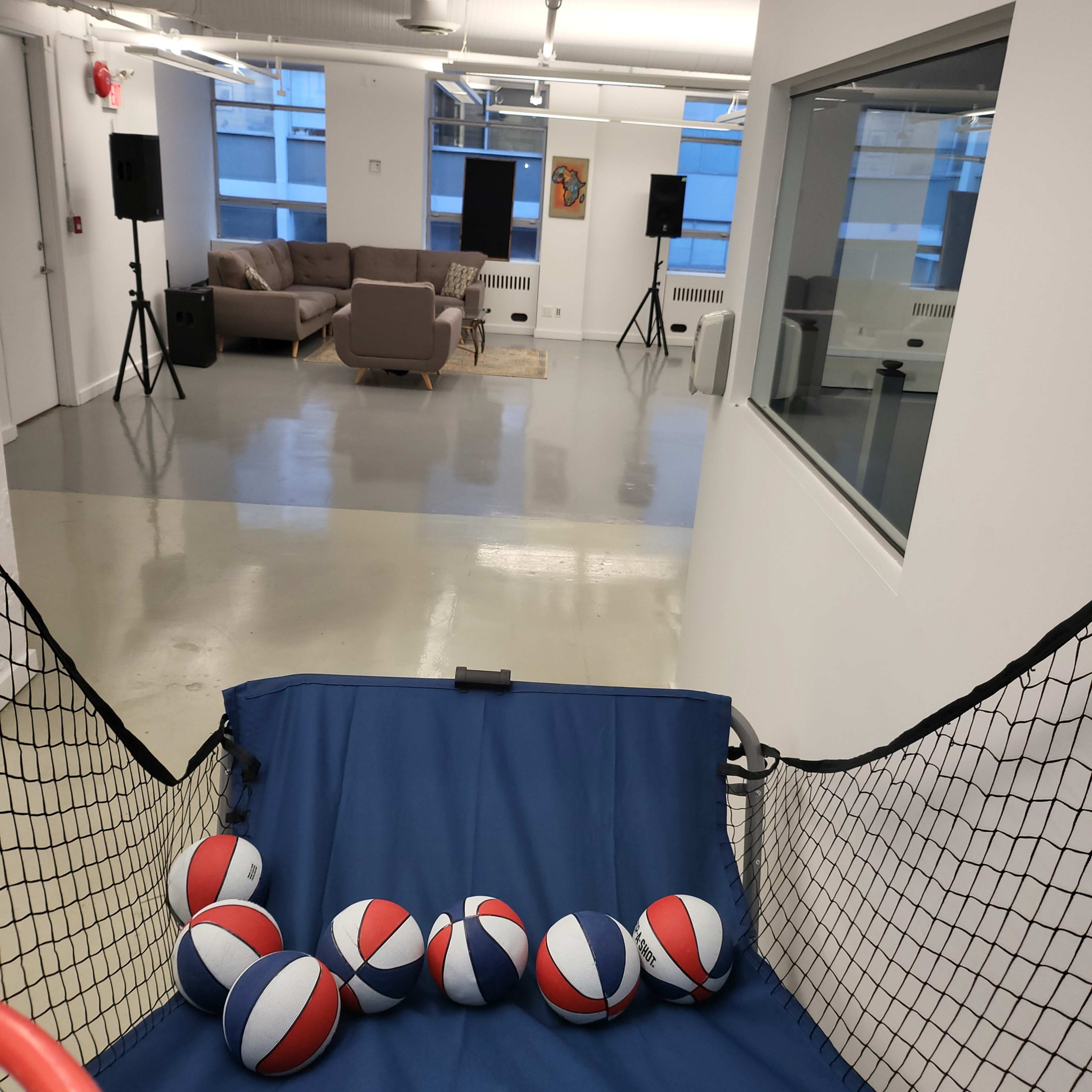 A spacious room with a basketball hoop and several basketballs in the foreground, leading to a lounge area with a couch and speakers in the background.