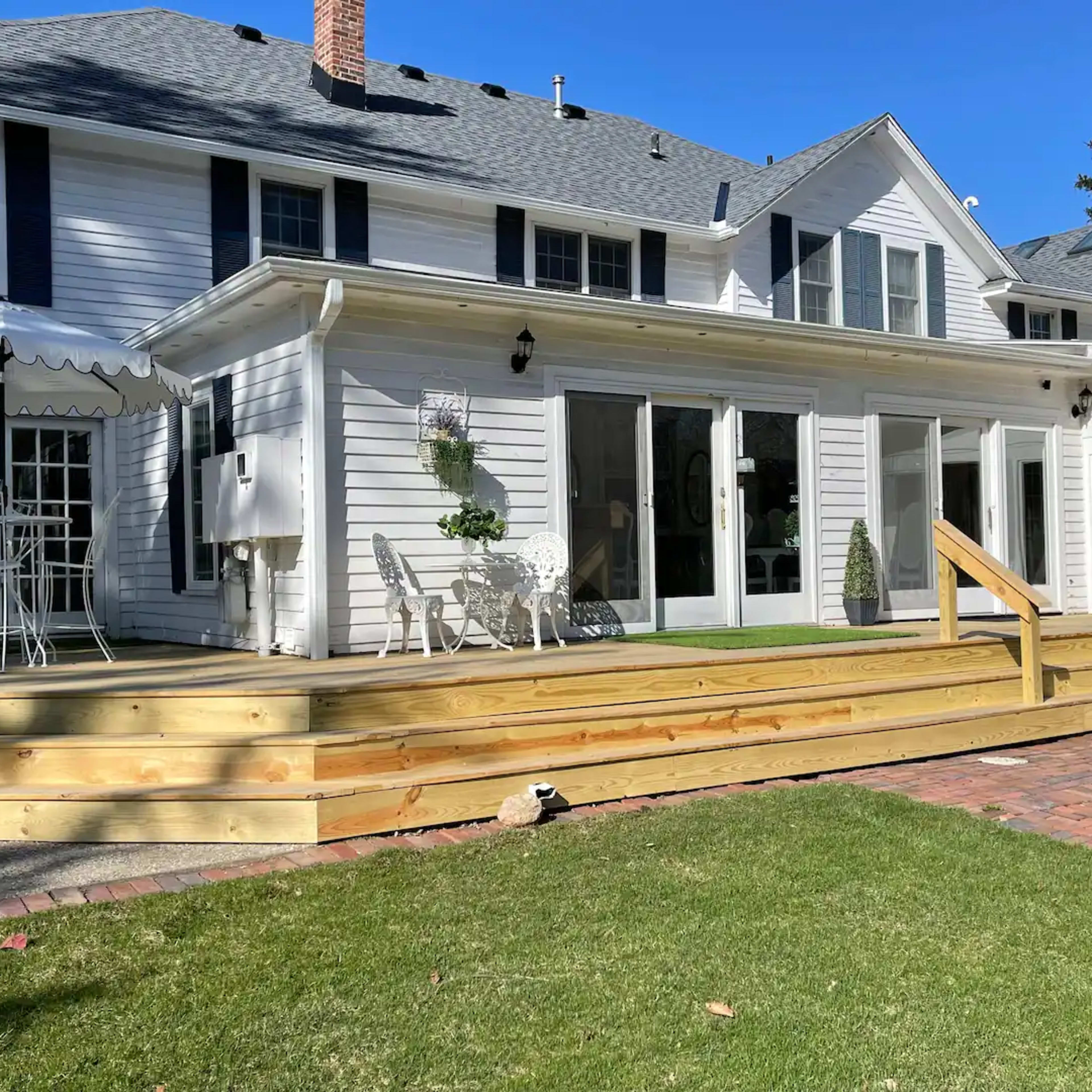 A two-story white house with a wooden deck, open patio furniture, and a landscaped lawn under a clear blue sky.