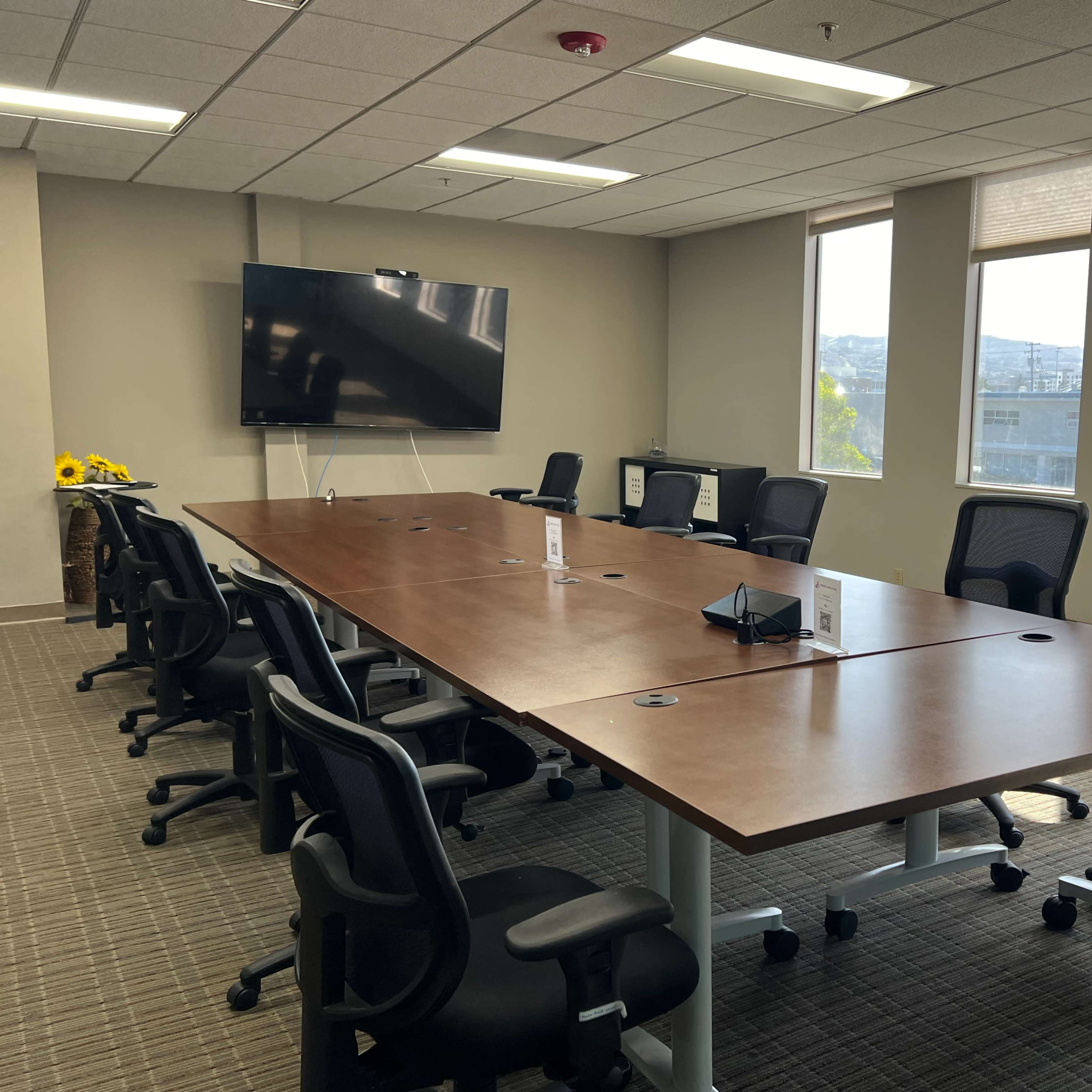 A spacious conference room features a large wooden table surrounded by black office chairs and a wall-mounted screen.