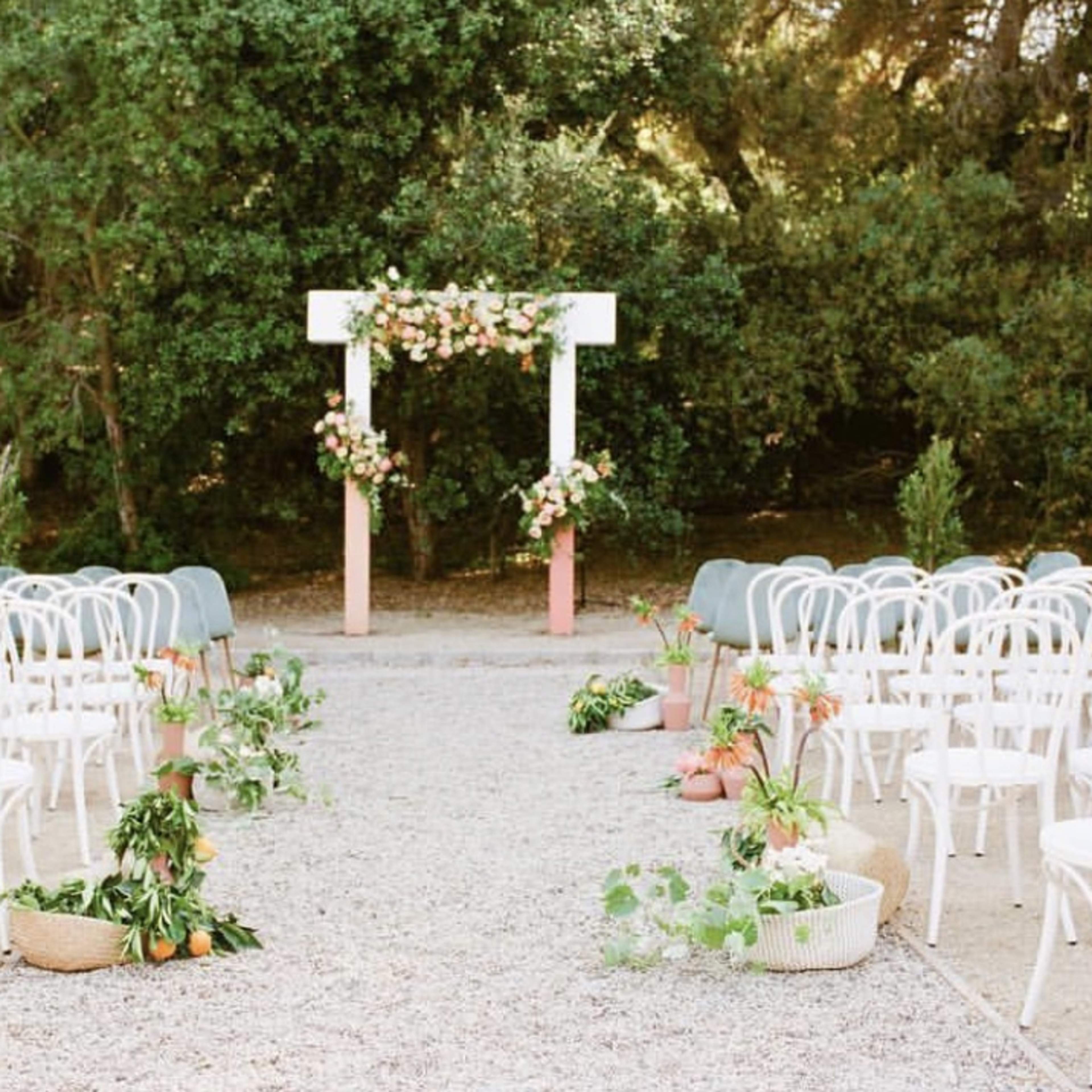 A wedding ceremony setup features white chairs arranged in rows facing a floral arch surrounded by greenery.