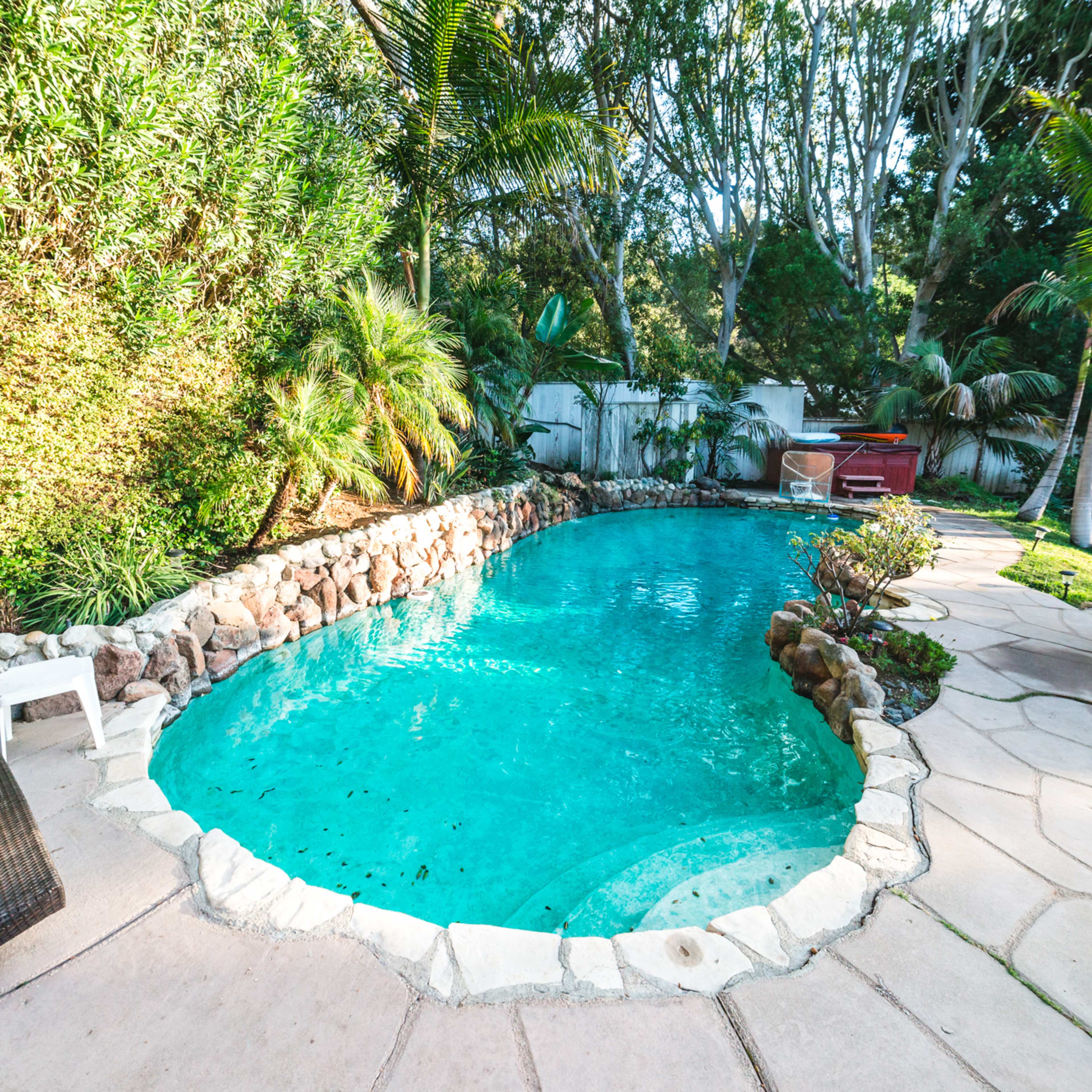 A curved pool surrounded by stone walls and tropical plants, with a paved walkway and a white chair nearby.