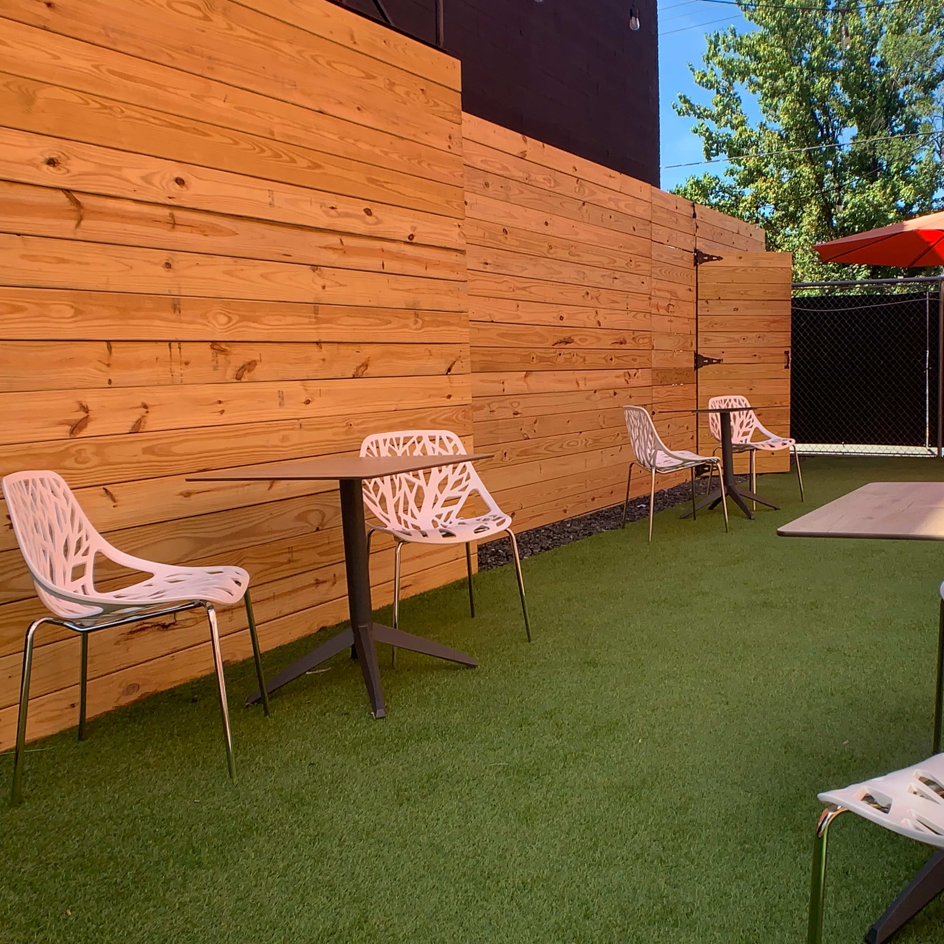 A small outdoor seating area with artificial turf, surrounded by wooden panels, featuring several chairs and tables, and an orange umbrella overhead.