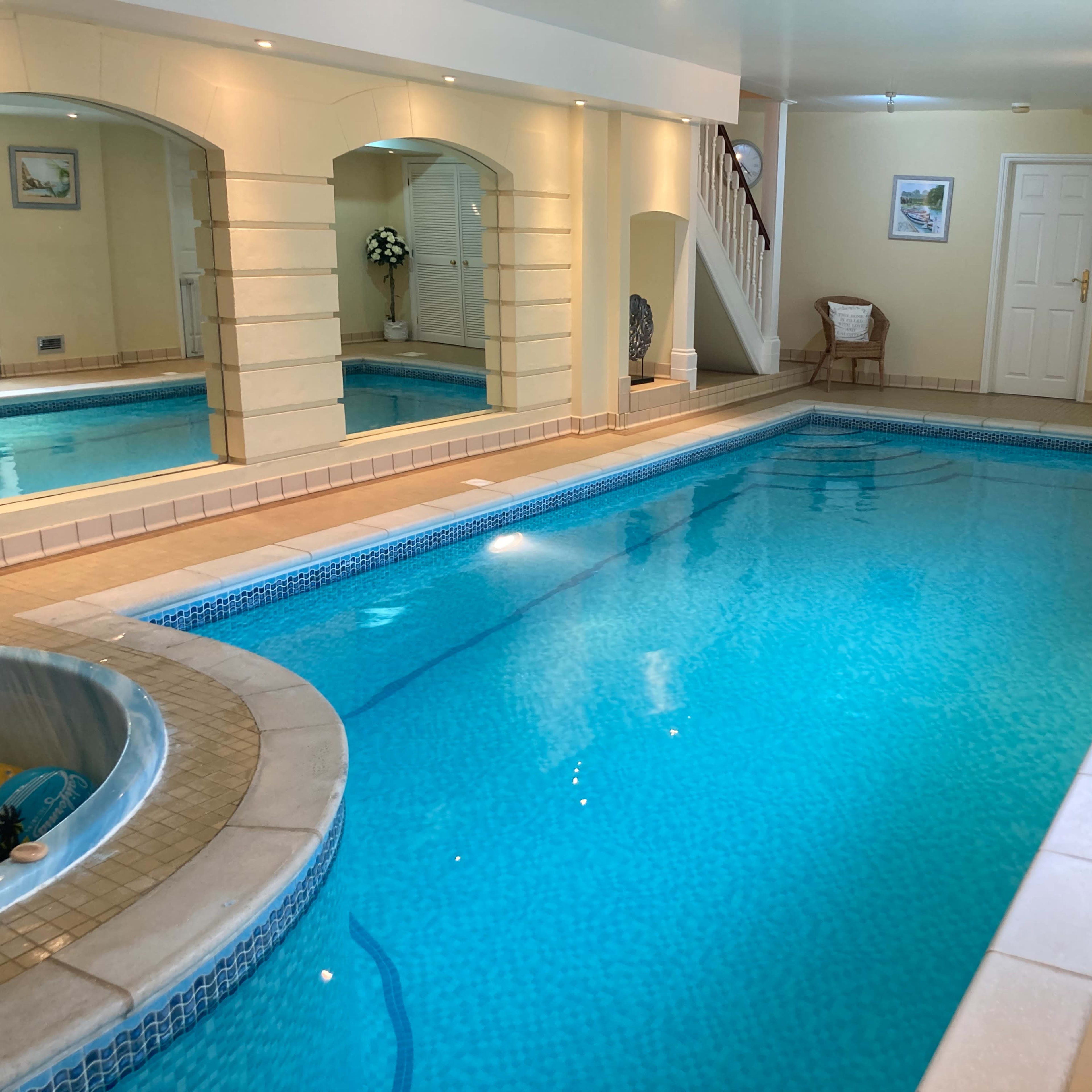 An indoor swimming pool area with blue water, surrounded by tiled walls and a jacuzzi on one side.