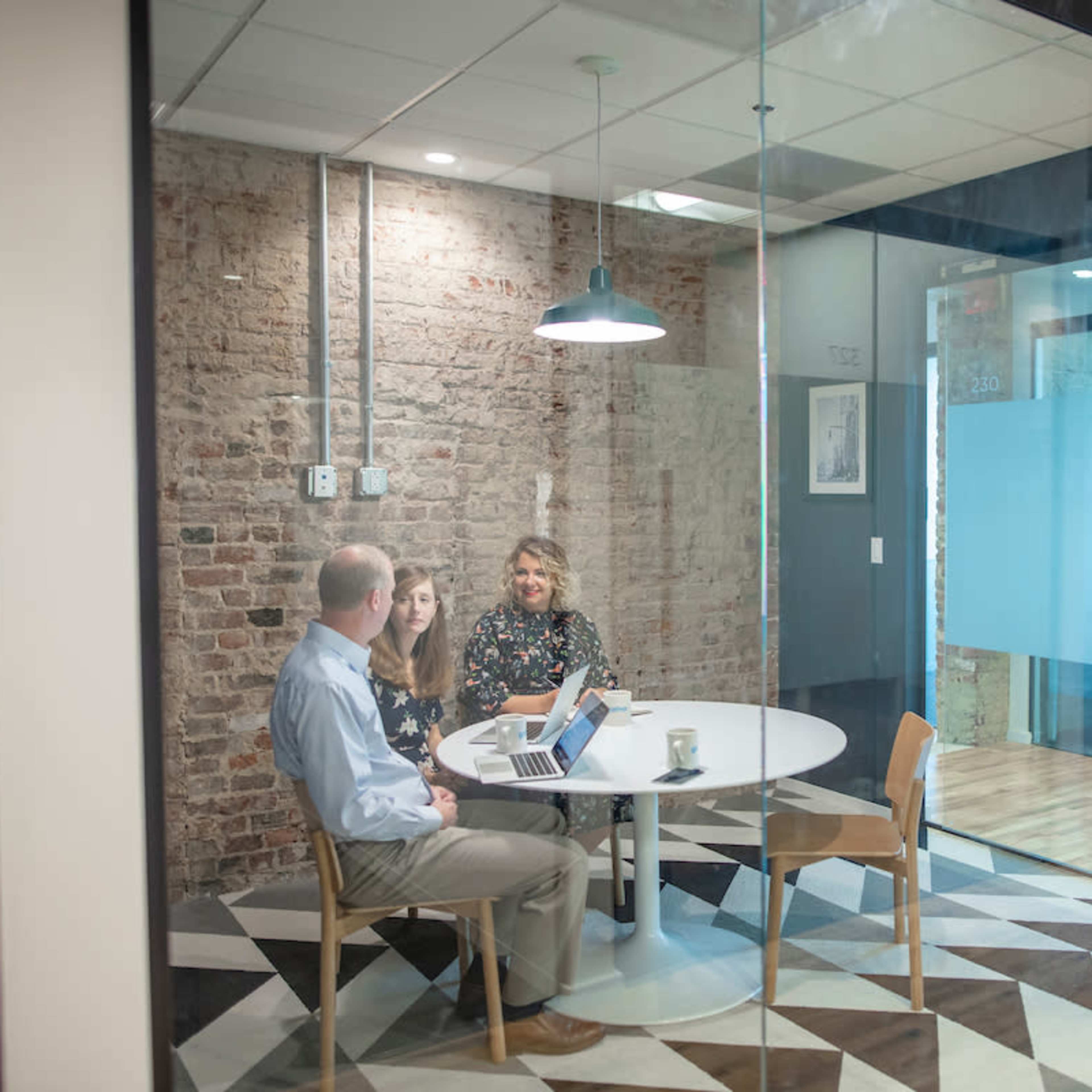 Three people are seated at a round table in a glass-walled conference room with exposed brick, discussing while using a laptop.