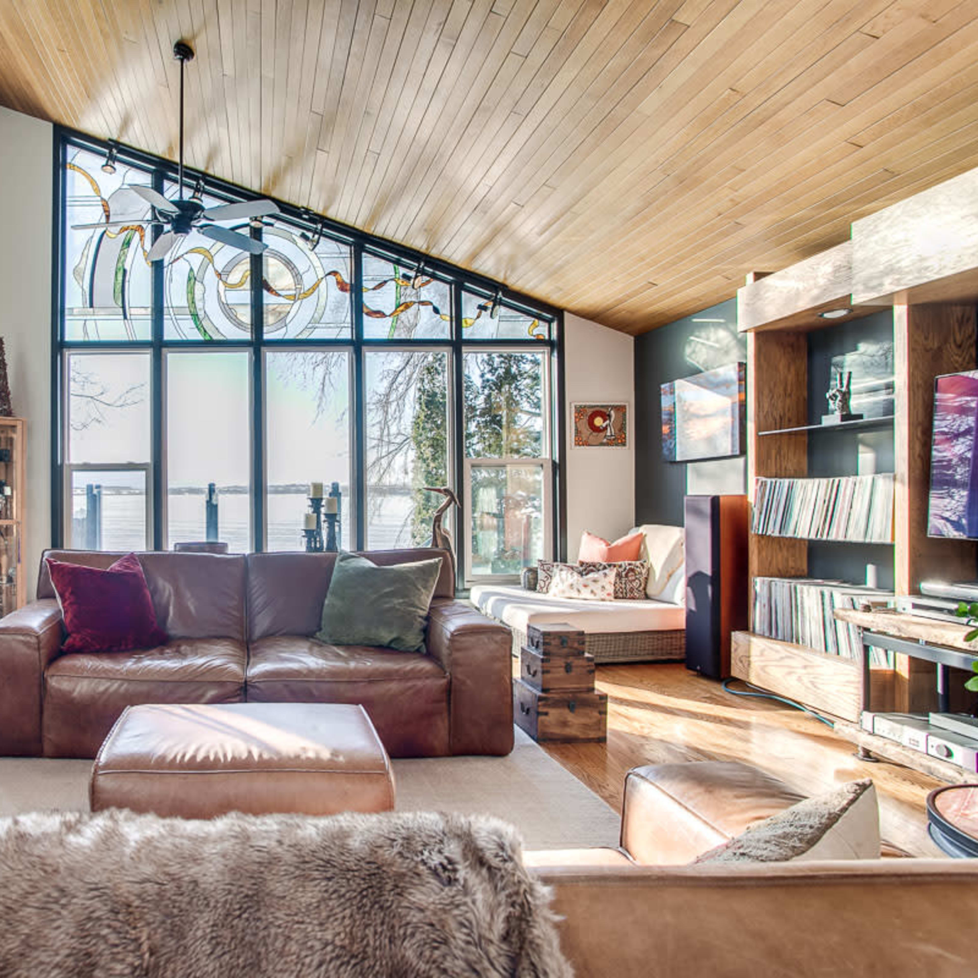 A spacious living room features large windows with a view of the water, wooden ceilings, and comfortable seating arrangements.